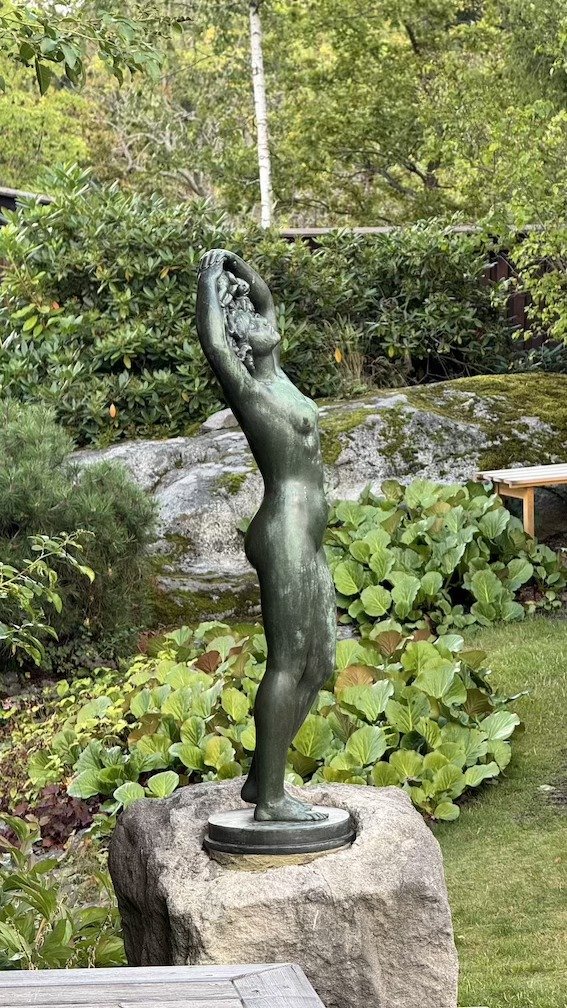 Bronze sculpture of a standing woman with arms raised, placed among greenery and stone in the museum garden.