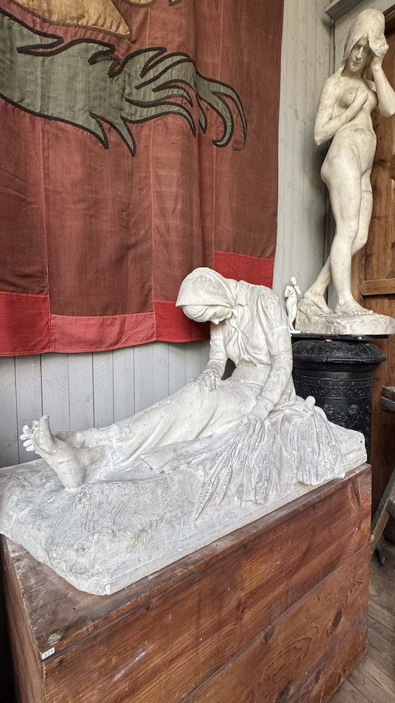 Plaster study of a reclining, mourning figure with head covered, lying beside a red banner and other plaster figures.