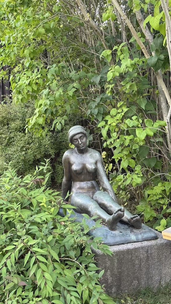 Seated bronze figure of a woman resting on a stone plinth, surrounded by dense green garden plants.
