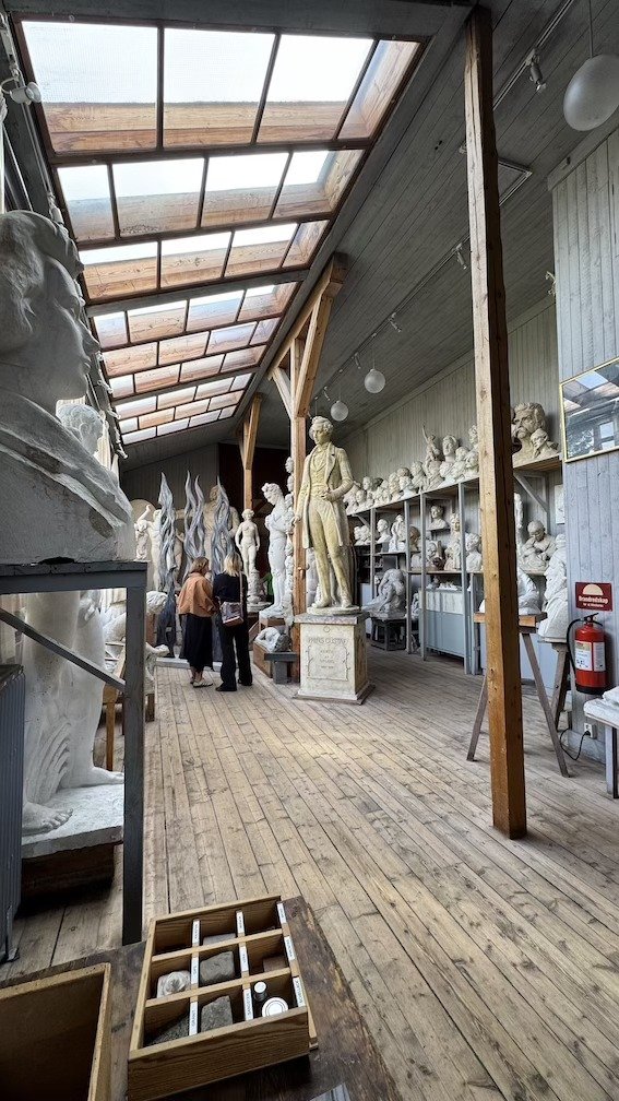 Long wooden gallery walkway under skylights, lined with rows of plaster portrait busts, with two visitors looking at the collection.