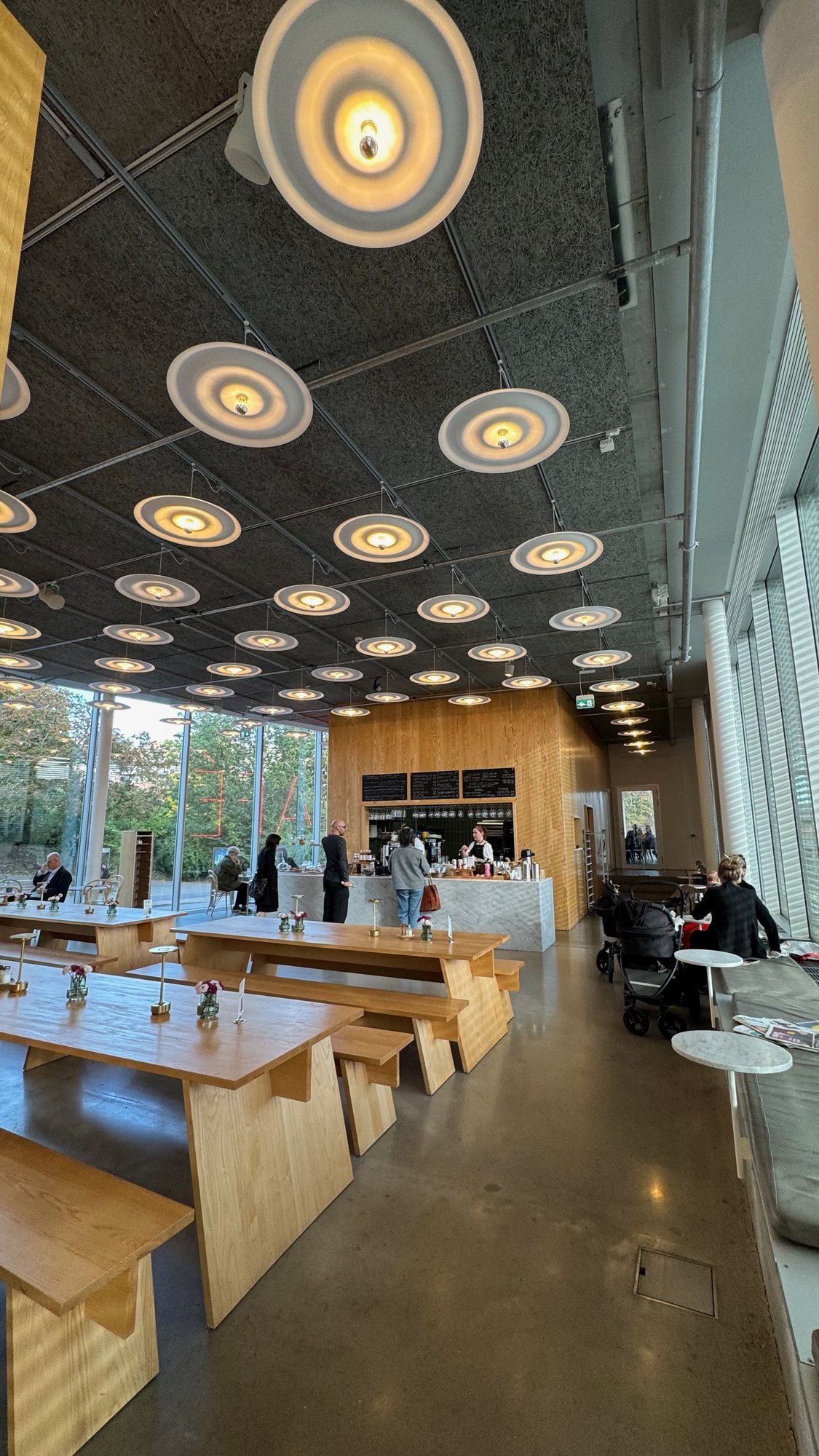 Café in Bonniers Konsthall with long wooden tables and benches, a wooden counter with a marble front, and round lamps hanging from a dark ceiling.