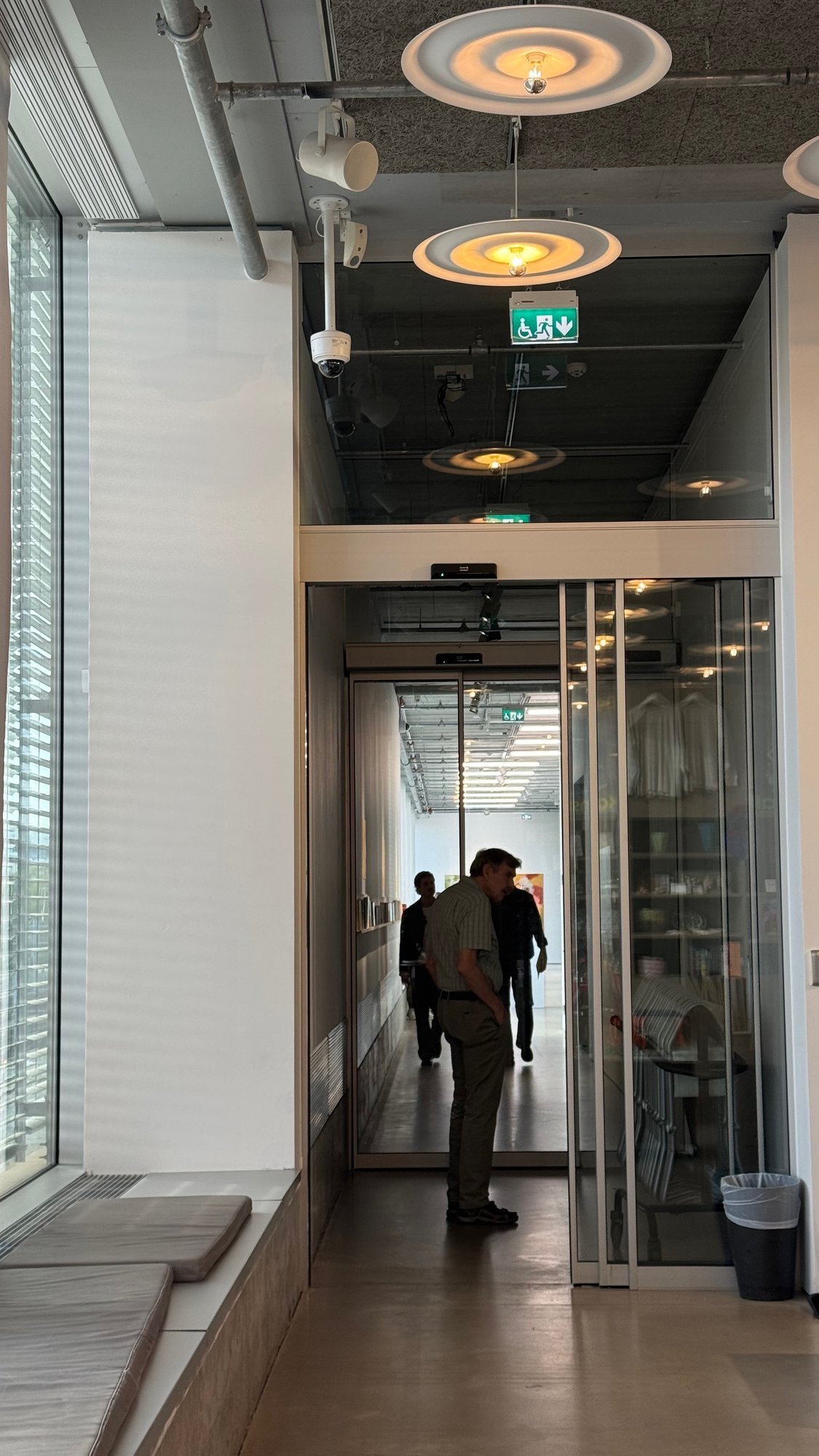 Corridor inside Bonniers Konsthall with round ceiling lamps and sliding glass doors. Visitors walk toward the galleries at the end of the corridor.