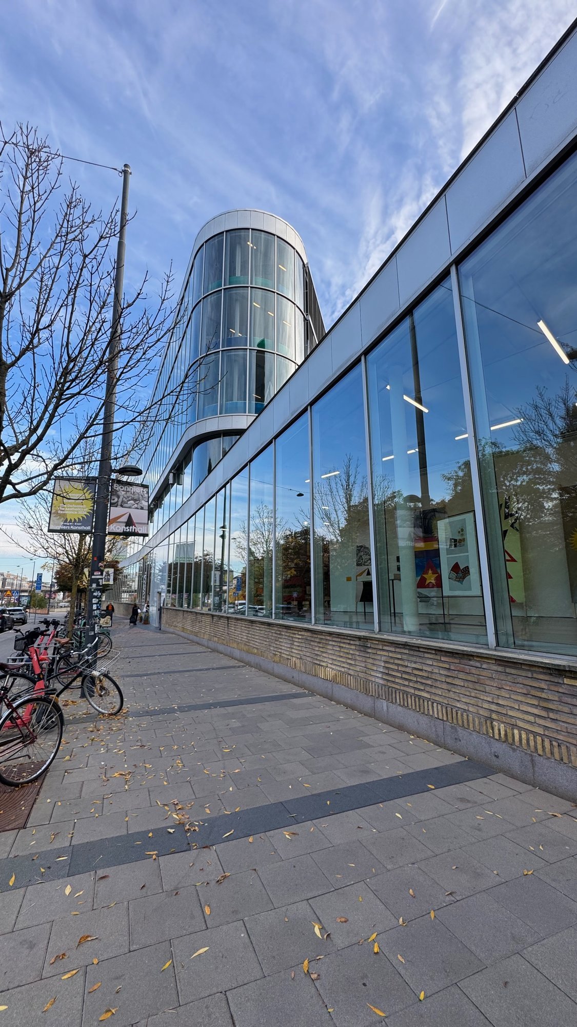 Long glass front of Bonniers Konsthall along a street, with a rounded glass tower behind it, banners on a lamp post and bicycles on the pavement.