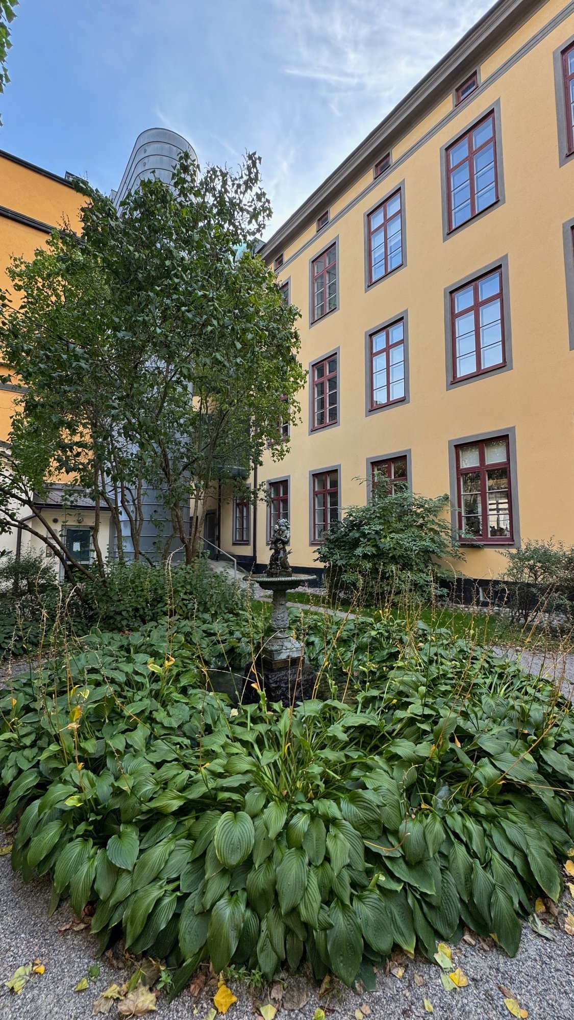 Courtyard of Almgrens Sidenväveri with a yellow building with red window frames, a small fountain in a bed of green hosta plants, and a grey tower behind the trees.