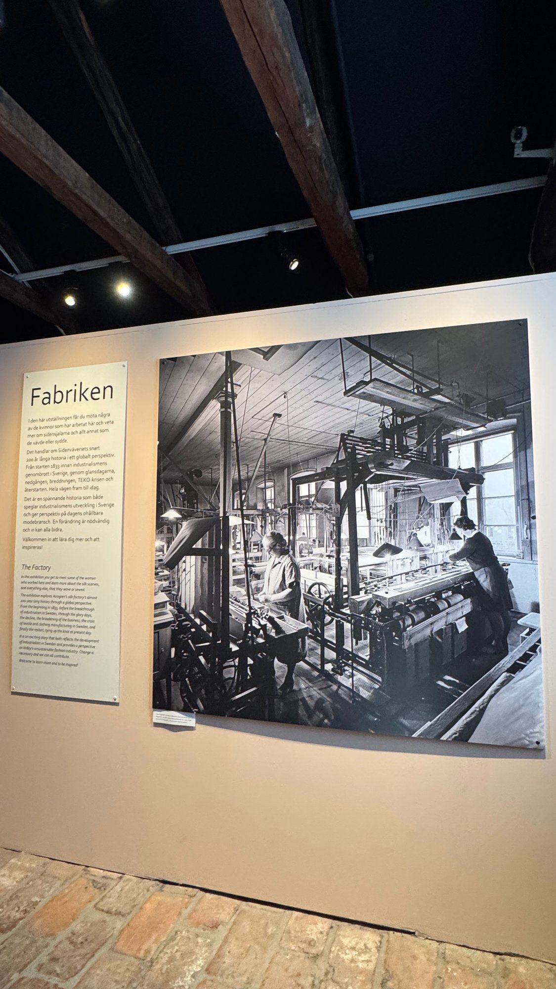 Wall in the Fabriken (The Factory) exhibition with a text panel in Swedish and English and a large black-and-white photo of two women working at looms in a factory hall.