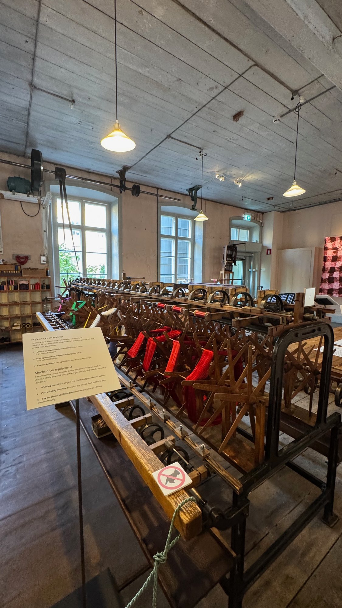 Silk winding machine with red silk skeins on wooden reels in the museum hall, with a sign titled Mekaniska maskiner (Mechanical equipment) and tall windows behind.