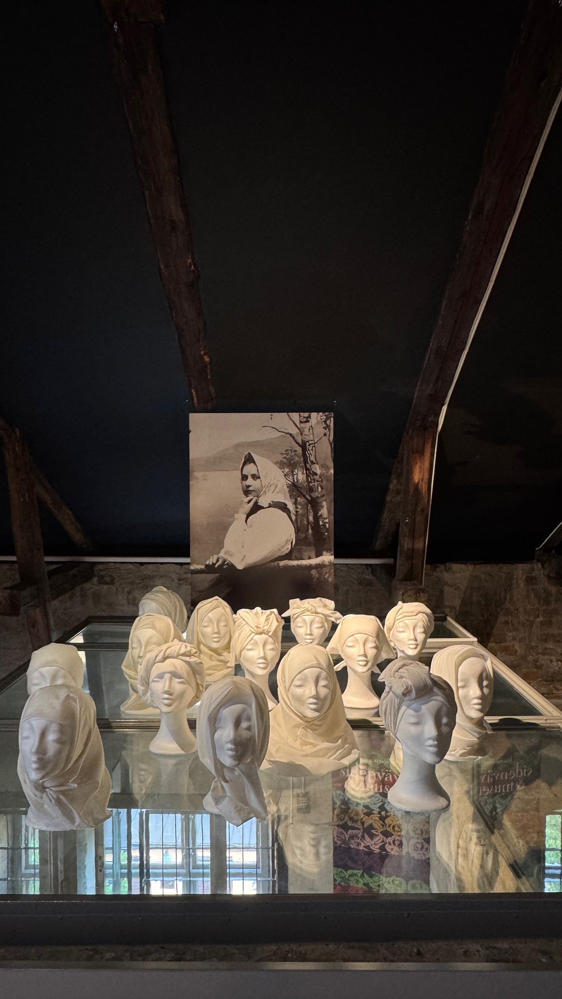 White plaster heads with scarves tied in different ways stand on a glass case. Behind them is an old black-and-white portrait of a woman in a headscarf.
