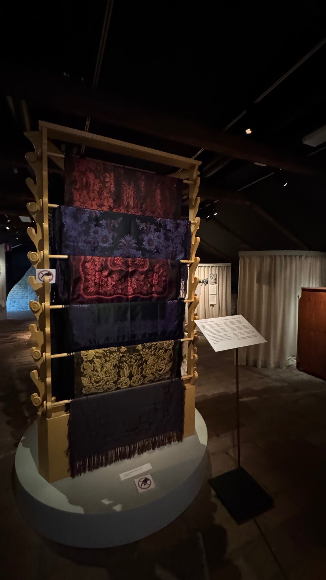 Six woven silk shawls with flower patterns in red, blue, green and gold hang on a wooden frame in a dim exhibition room.