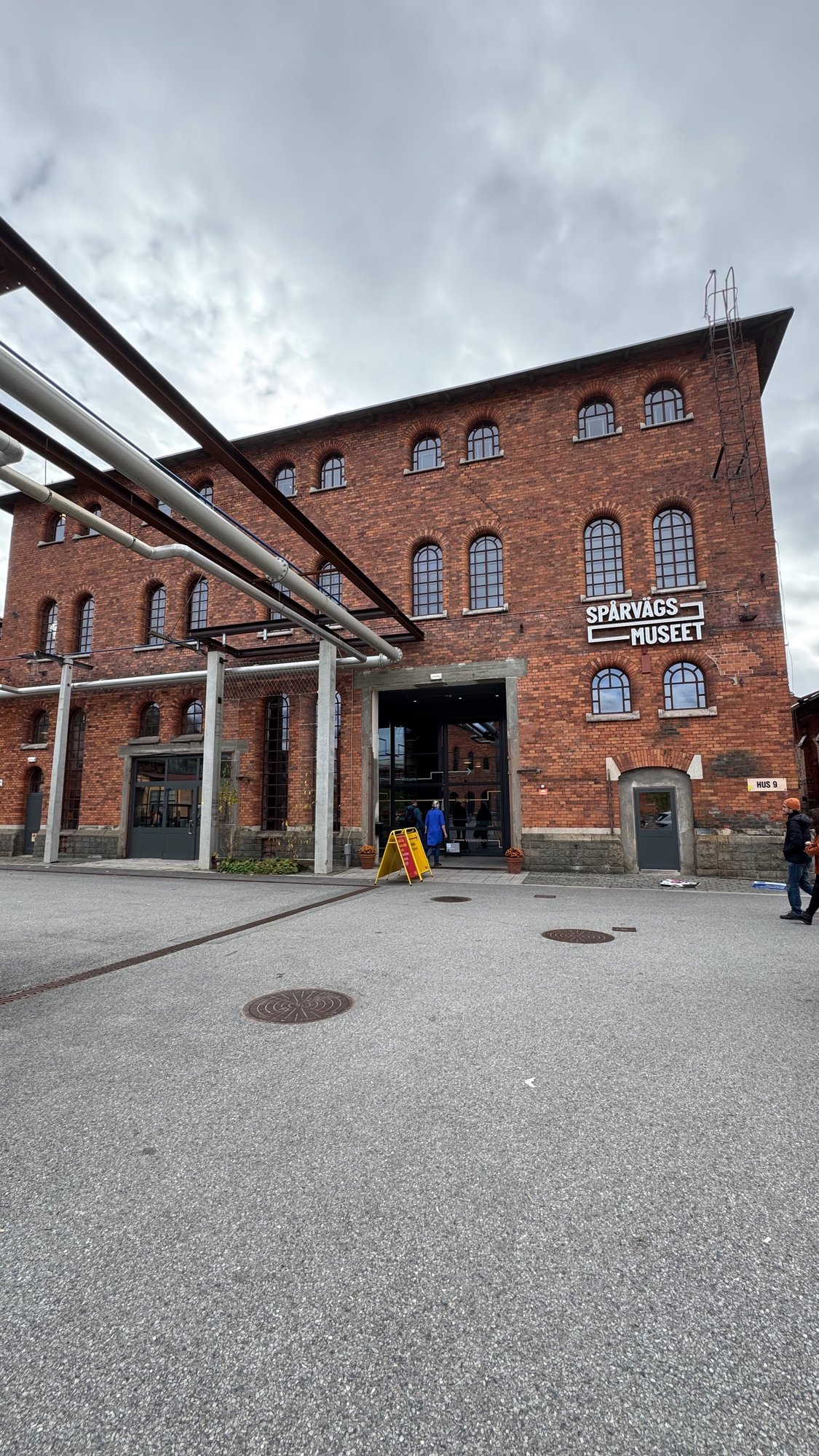 Red brick building with arched windows and the sign Spårvägs Museet above a wide glass entrance. A small sign on the right says Hus 9. Steel pipes run along the left side.