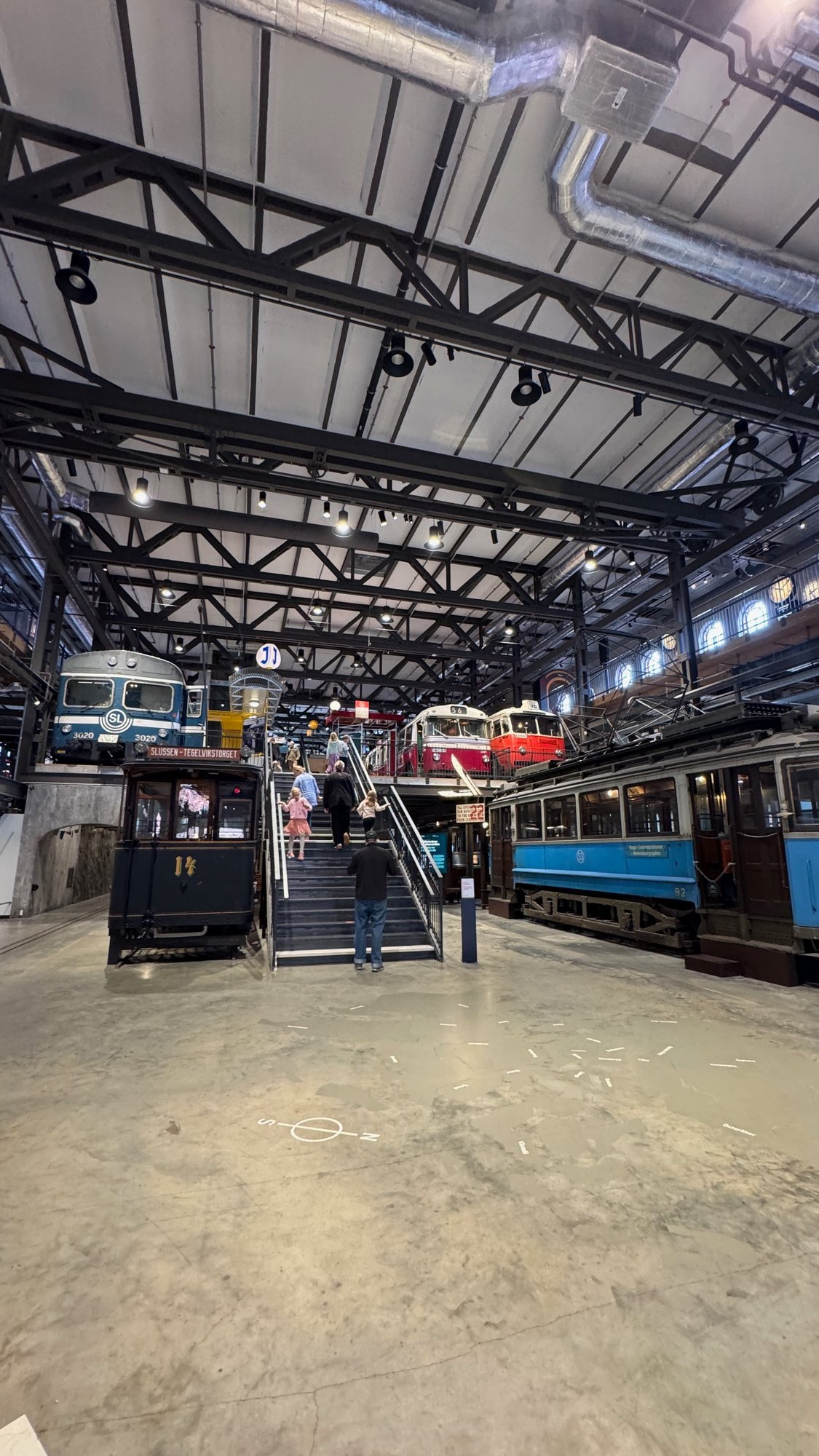 Wide view of the museum hall with a black tram with the number 14, a blue SL train, a red bus, a red and white bus and a blue tram under black steel roof beams. Visitors walk up a staircase.