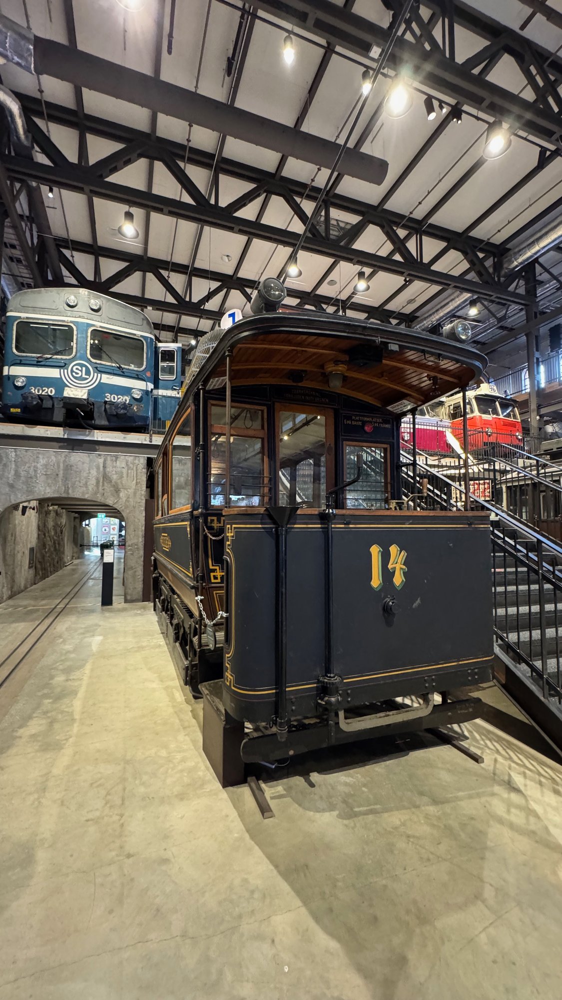 Dark blue tram with gold lines and the number 14, a destination sign for Slussen–Tegelviktorget, an open rear platform and a wooden roof. A blue SL train and a stone arch are behind it.