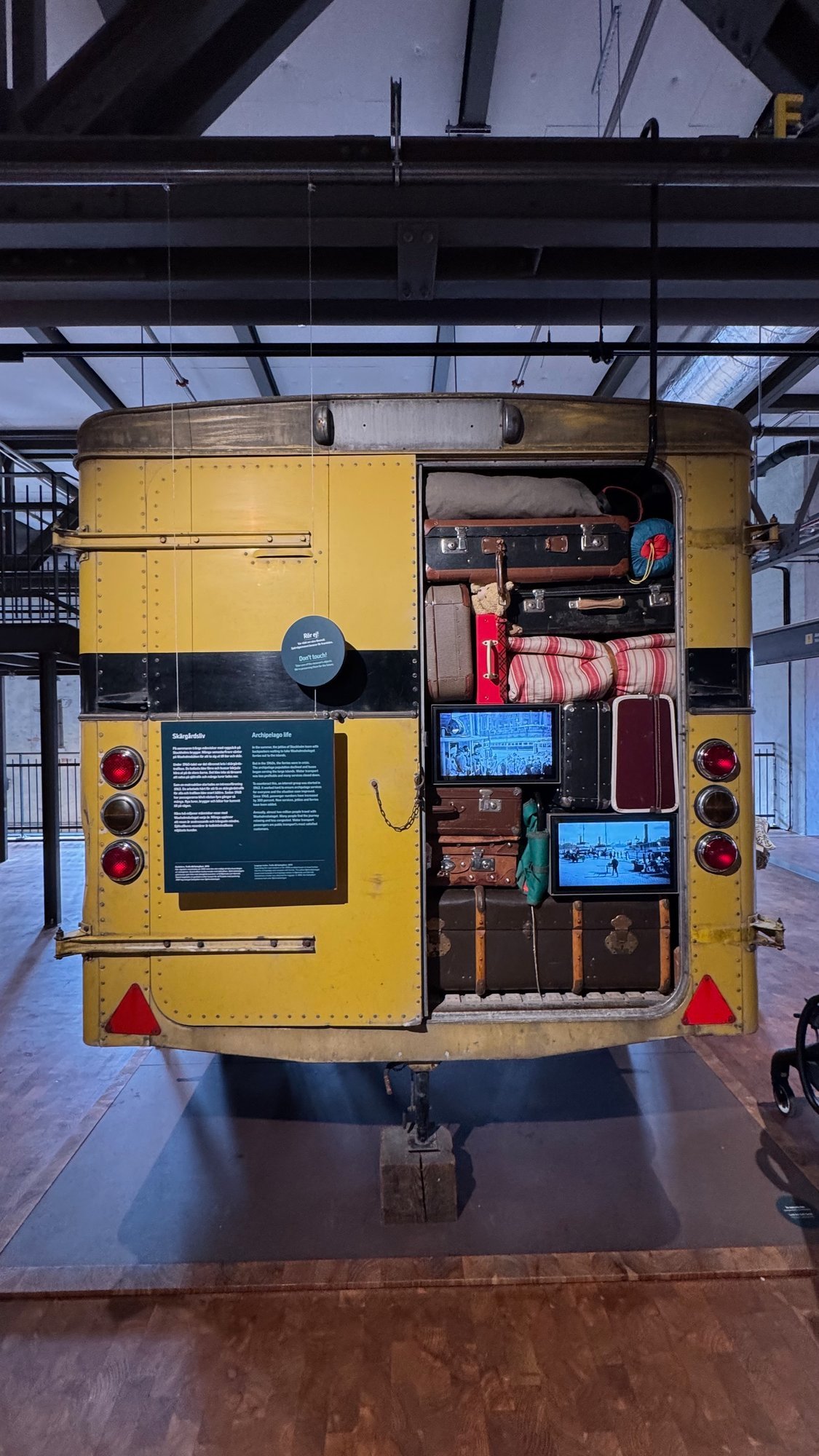 Rear of a yellow bus with an open luggage compartment full of old suitcases, a blanket and two small screens. A panel in Swedish and English titled Skärgårdsliv, Archipelago life, hangs on the left door.