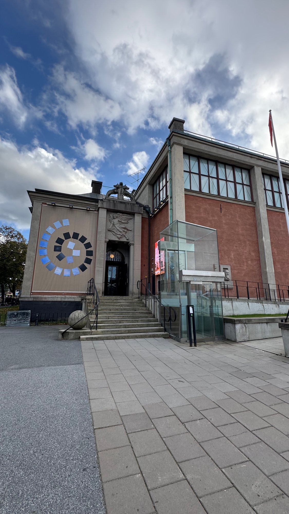 Liljevalchs art museum seen from the square: stone steps to a doorway with a carved winged figure, a banner with blue and black squares in a spiral on the left, red-brown walls with tall windows, and a glass lift on the right.