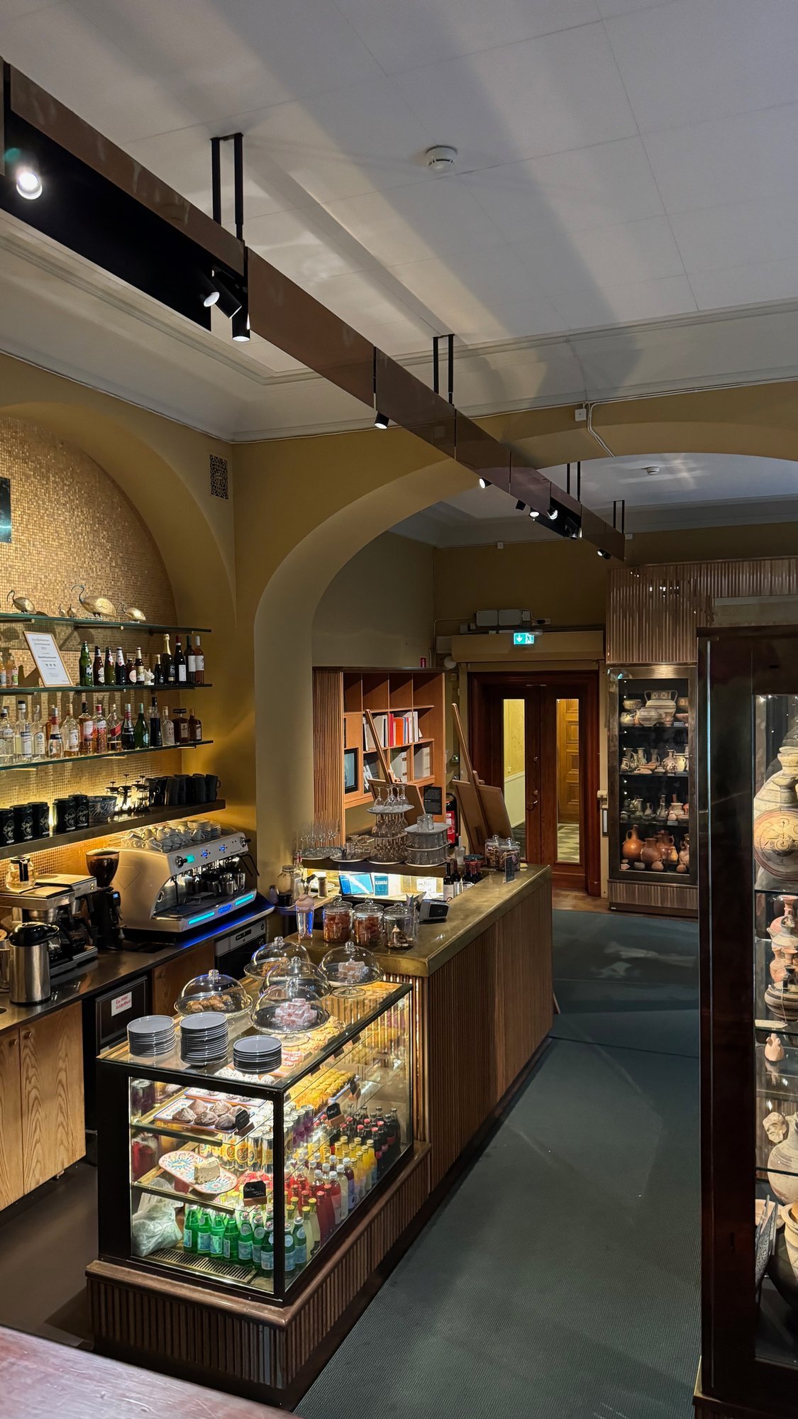 The counter of Bagdad Café in Medelhavsmuseet: a coffee machine, a glass display with pastries and drinks, bottles on shelves, a bookcase, and a display case with pottery on the right.