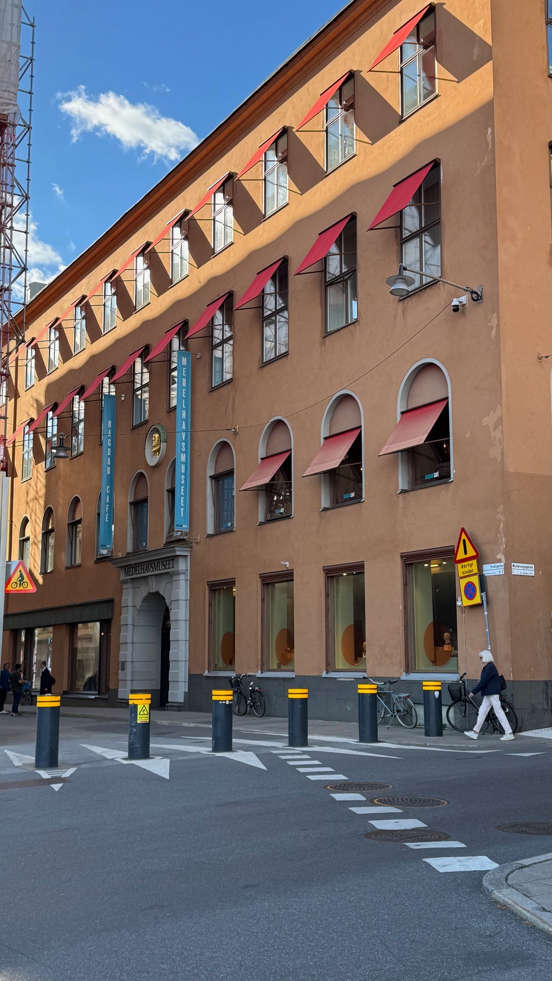 The orange facade of Medelhavsmuseet seen from the street corner, with red awnings, arched windows, two blue banners and black bollards on the street.