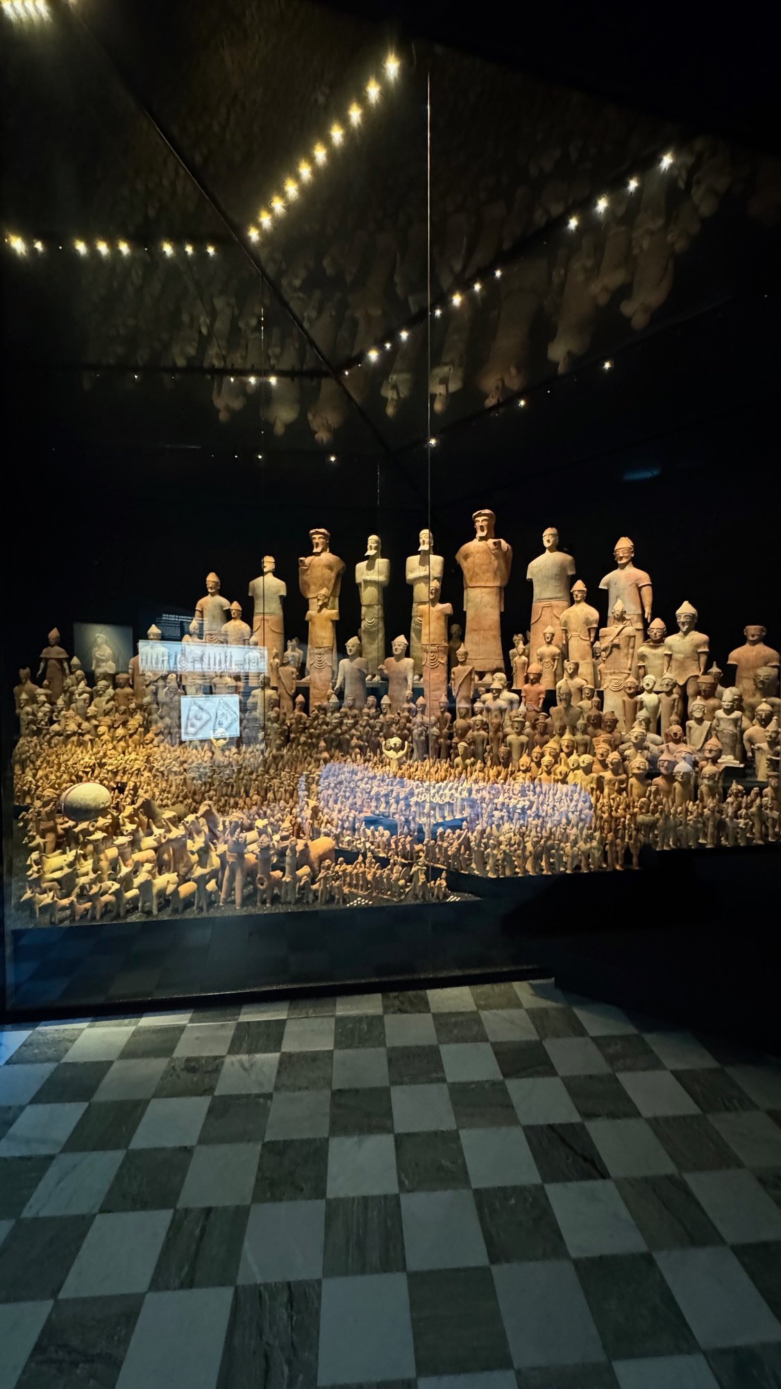 A large glass case with hundreds of clay figures, from tall standing figures at the back to small figures at the front, and clay animals on the left, in a dark room with a chequered floor.