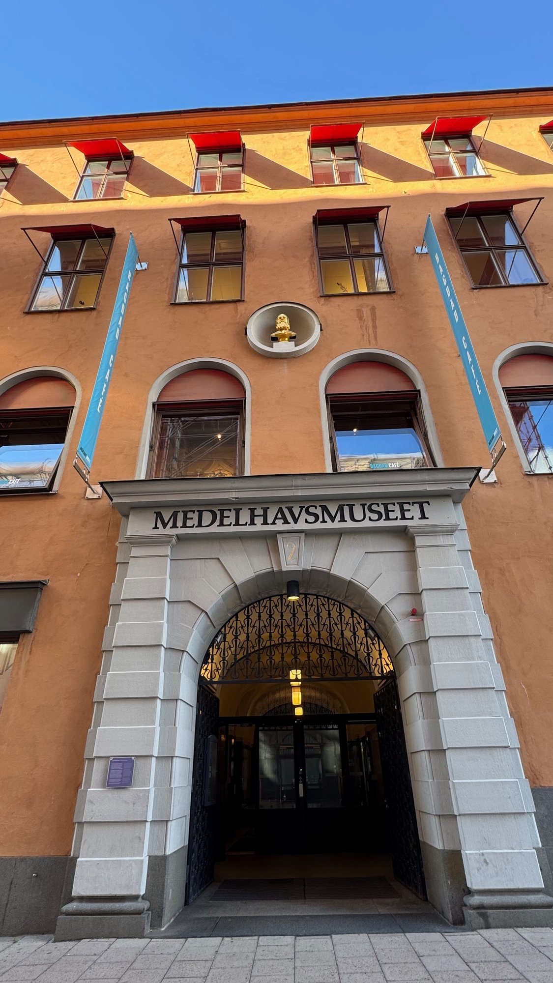 Front entrance of Medelhavsmuseet: a stone portal with the museum name above an arched wrought-iron gate, in an orange building with red awnings and a small golden bust in a round niche.