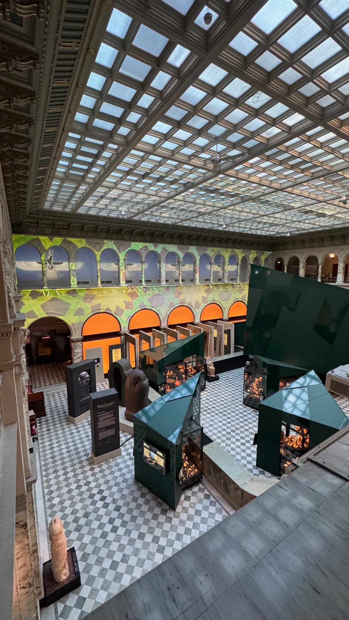 View from an upper floor into the hall of Medelhavsmuseet: a glass roof, a chequered marble floor, green angular display cases, a dark stone sarcophagus and a wall with a projected pattern.