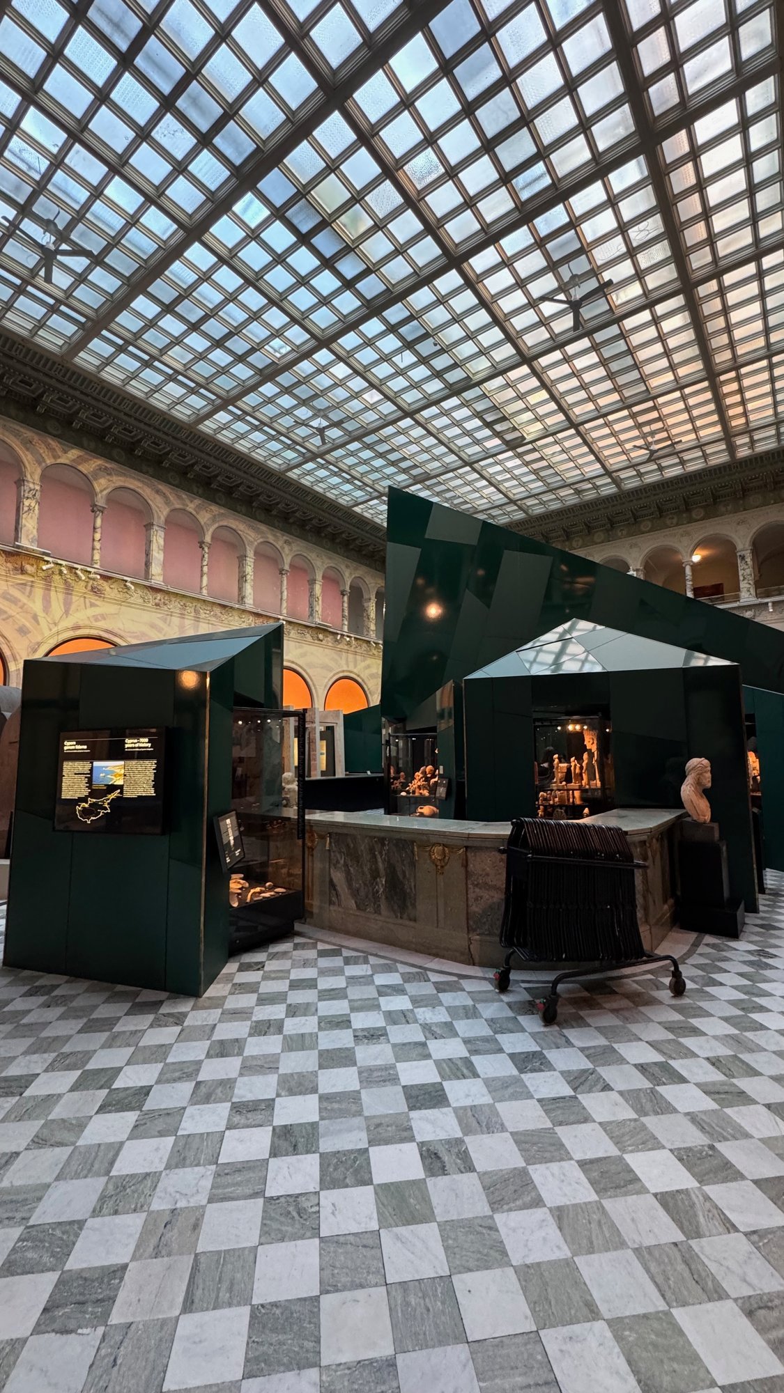 Green angular display cases with a sign for the Cyprus exhibition under the glass roof of the hall, on a chequered marble floor, with a marble bust on the right.