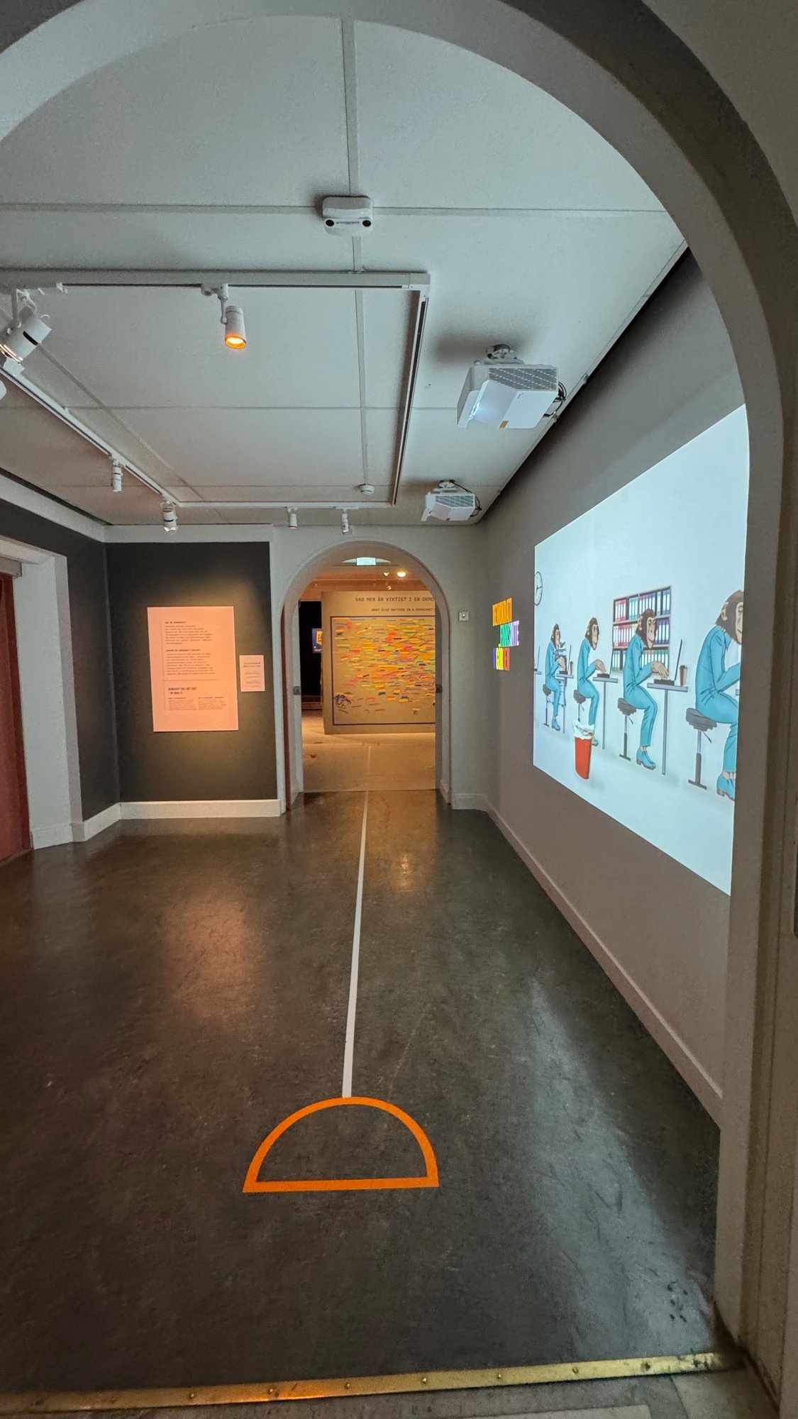 A corridor with a projected animation of apes in blue suits working at desks, text panels on the left and a wall of coloured notes in the next room.