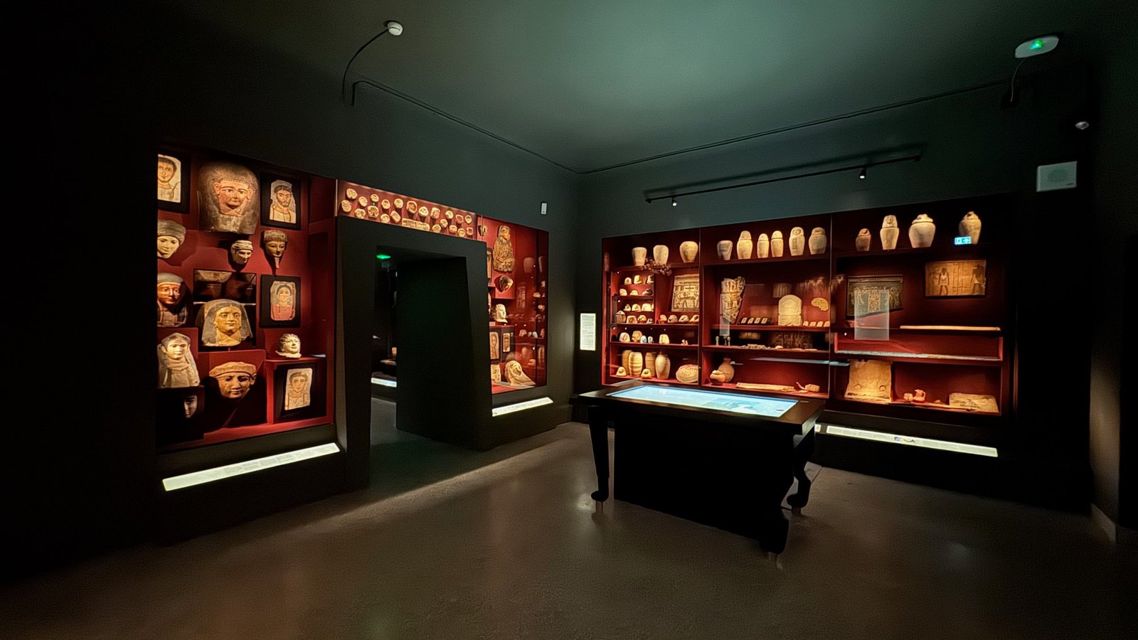 A dark exhibition room with red display walls: painted portrait faces and masks on the left, jars, tablets and small objects on the right, and a table with a blue touch screen in the middle.