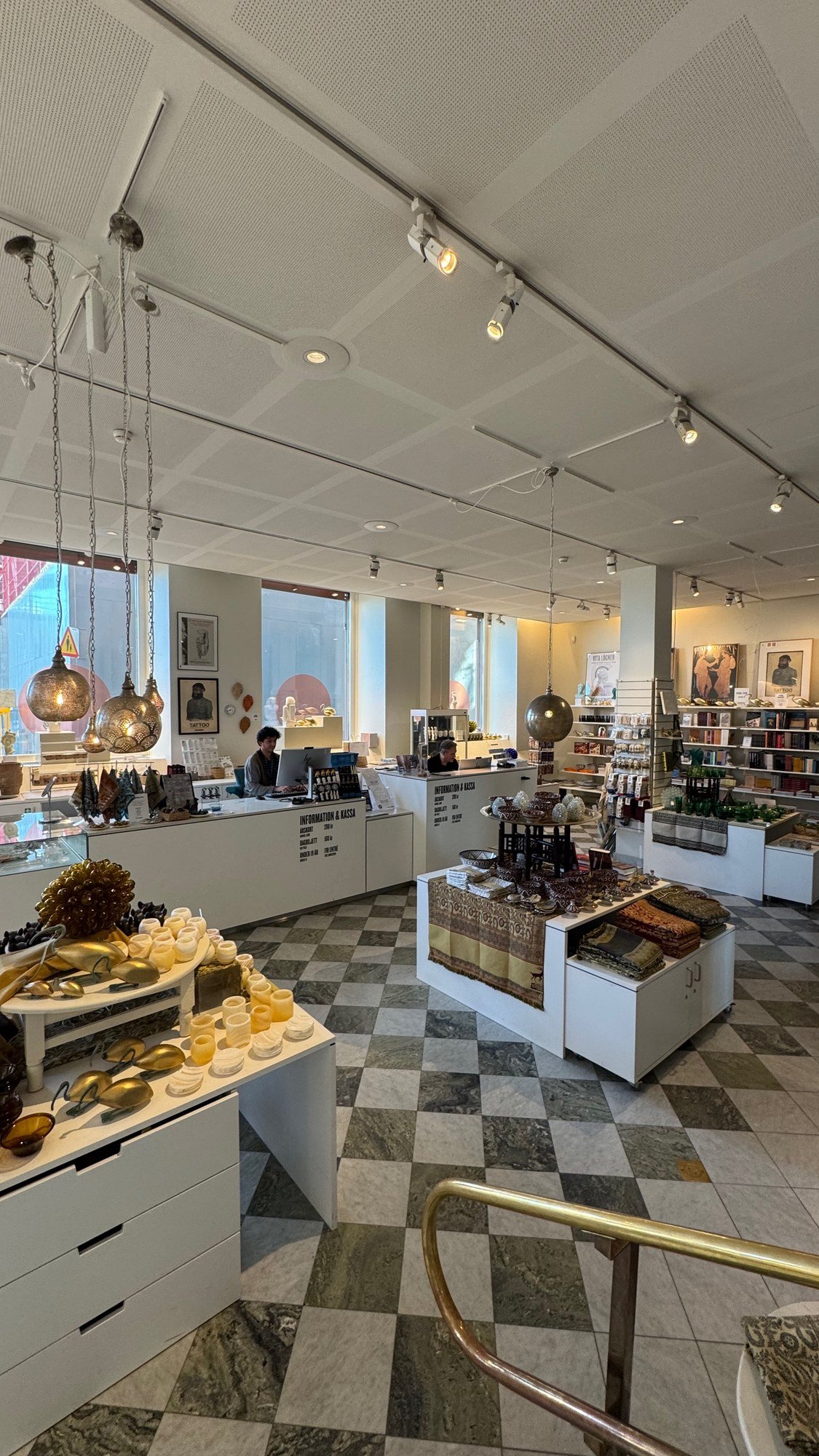 The museum shop at Medelhavsmuseet: white display tables with hanging pierced-metal lamps, candle holders, scarves and ceramics, a white information and cash desk with two people behind it, bookshelves on the right and a chequered marble floor.