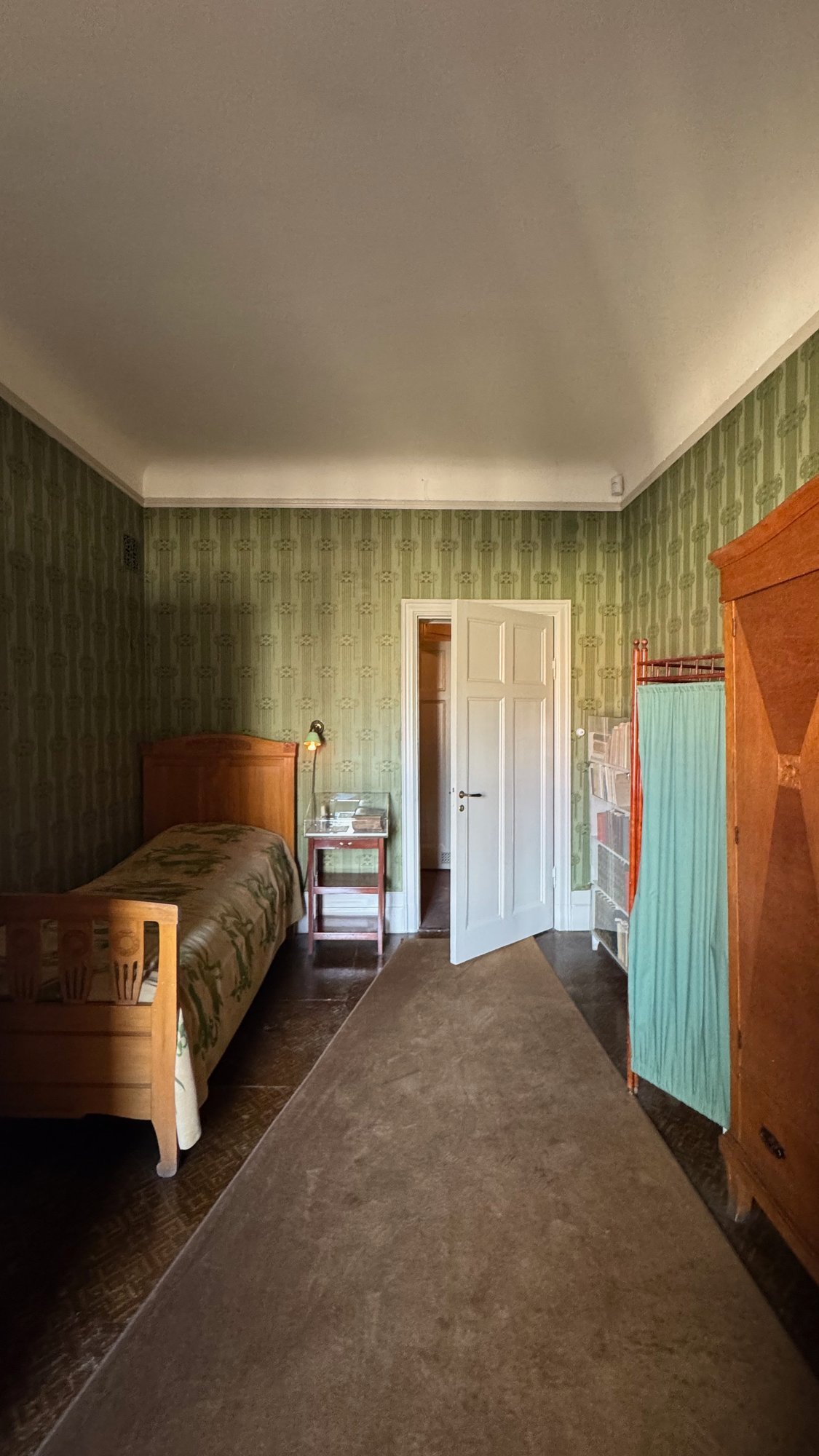 Narrow bed in a room with green patterned wallpaper, with a view through an open door to the next room.