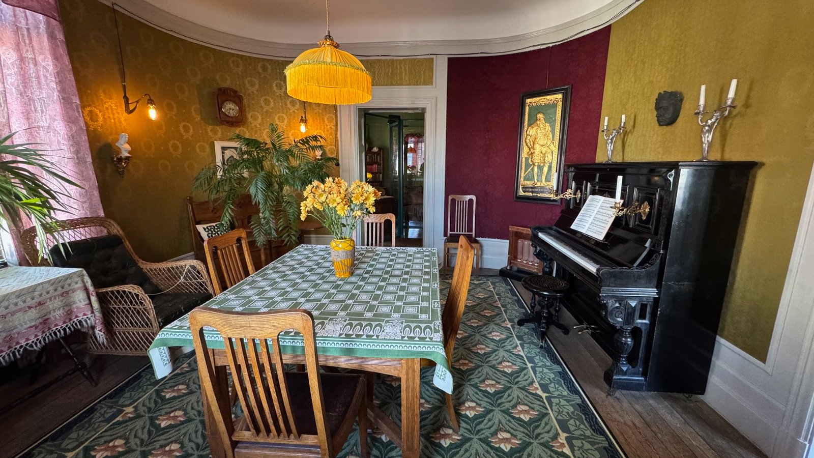 Dining room with a laid table with a green checked cloth, wooden chairs, a black piano and red and yellow wallpaper.