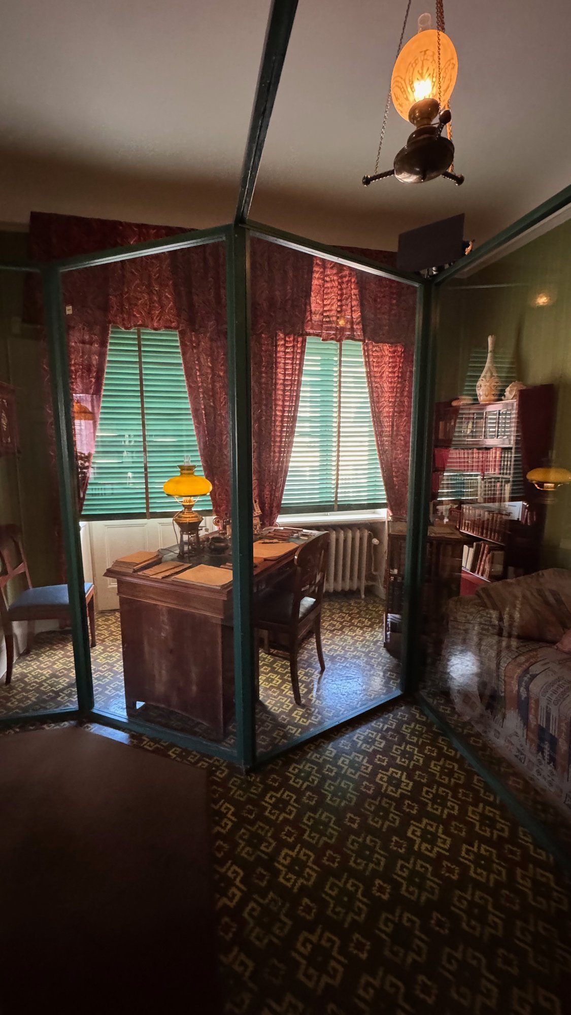 Writing desk and chair behind glass walls in a room with red curtains and green window blinds.