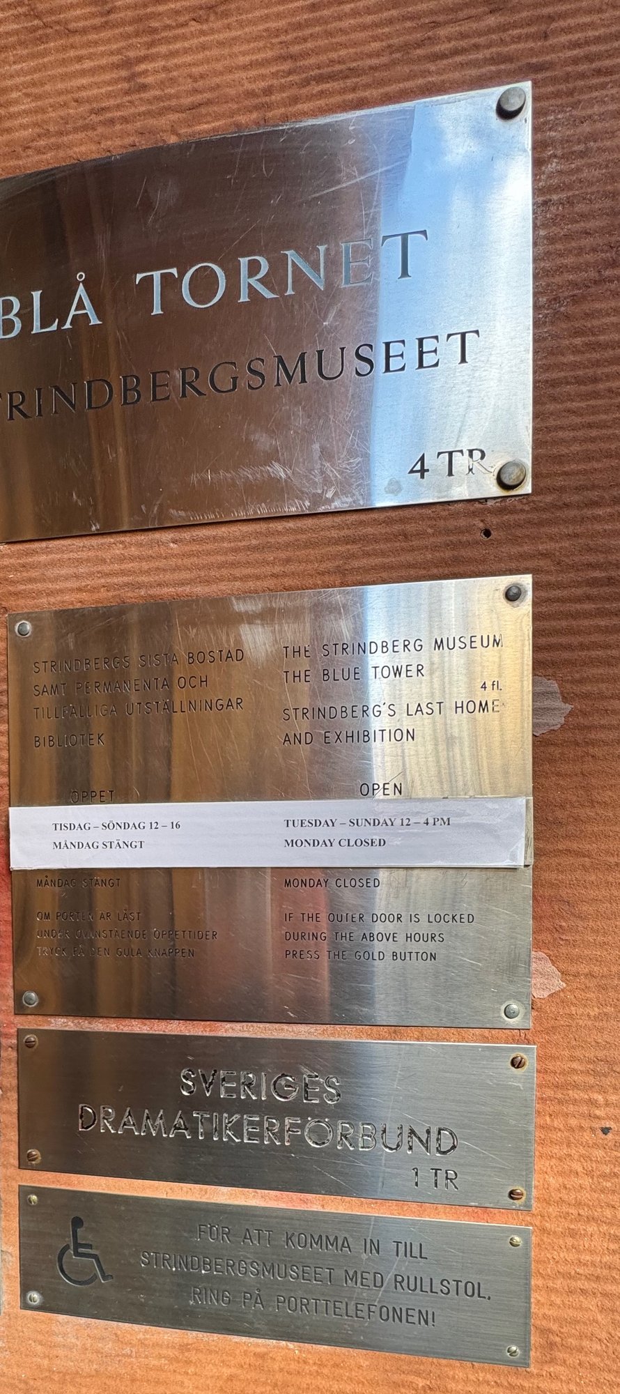 Metal signs beside the entrance door: Blå tornet and Strindbergsmuseet 4 tr, opening hours Tuesday to Sunday 12 to 4 pm with Monday closed in Swedish and English, a sign for Sveriges Dramatikerförbund, and a wheelchair sign that says to ring the intercom.