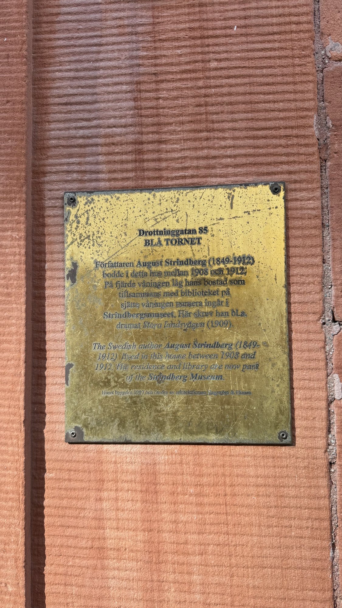 Brass plaque on a red-brown stone wall with the title Drottninggatan 85, Blå tornet, and text in Swedish and English about August Strindberg living in the house between 1908 and 1912.
