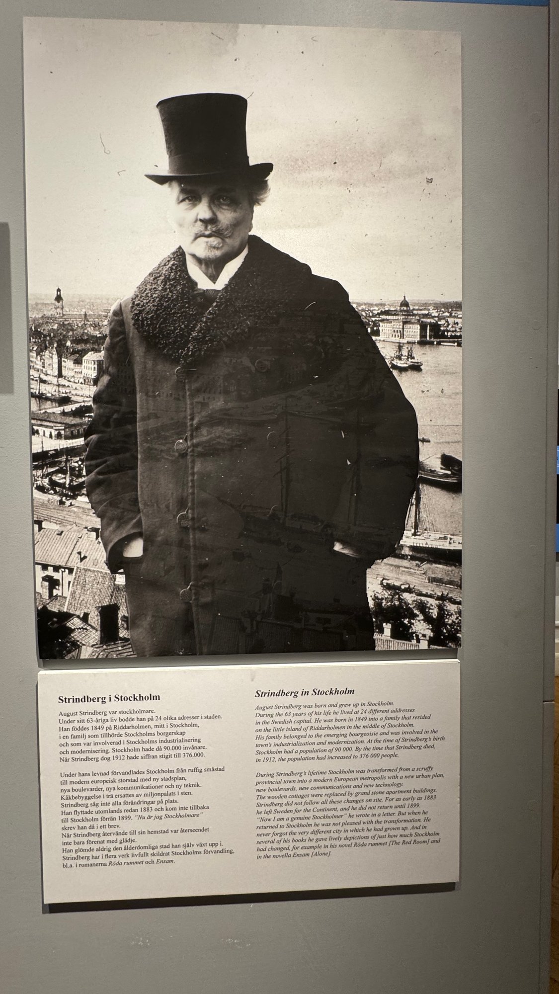 Large black and white photo of a man in a top hat and long coat on a wall, with a text panel below it.