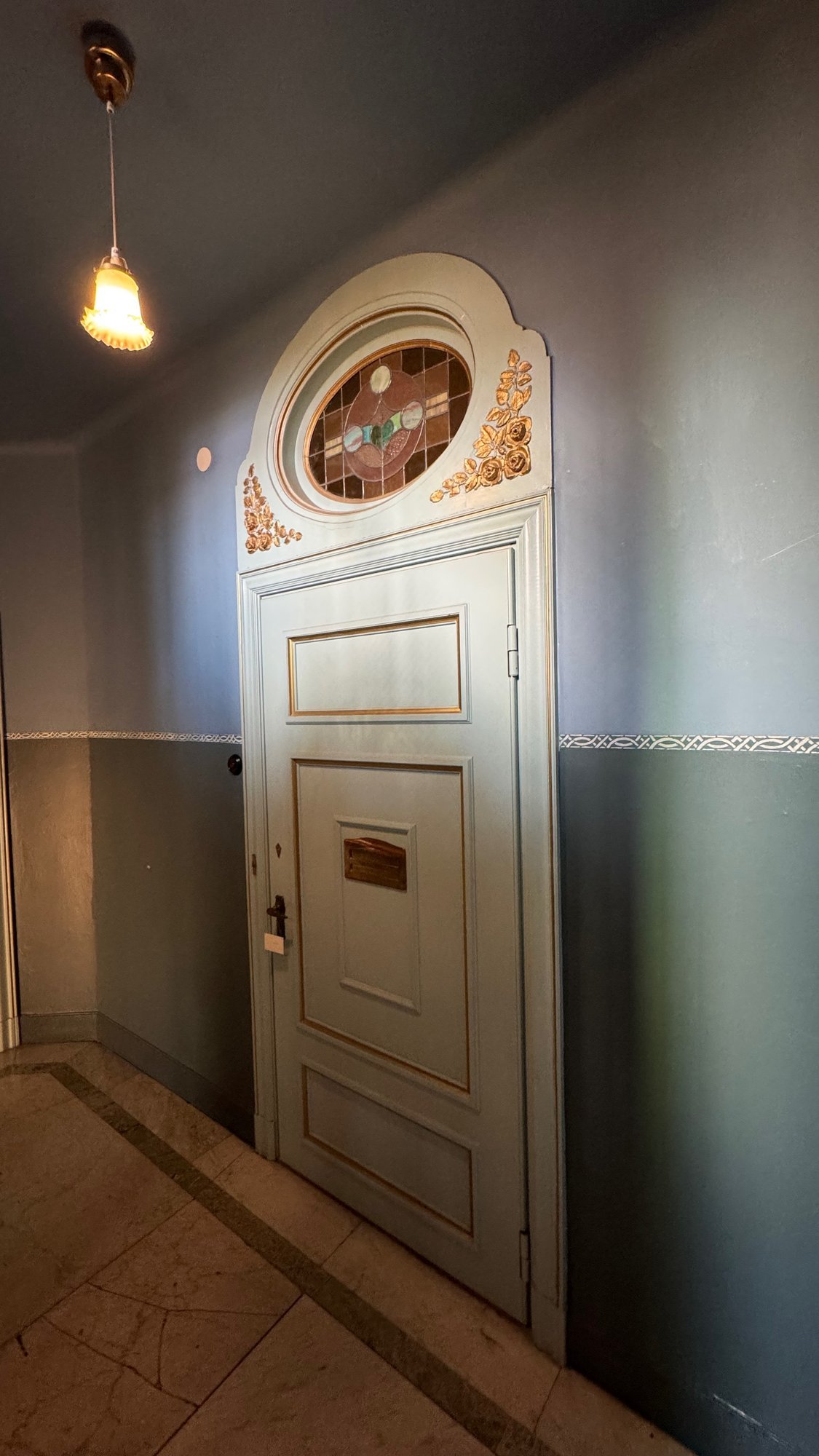Pale green door with gold edging and gold roses, and an oval stained glass window above it, in a blue-grey corridor.