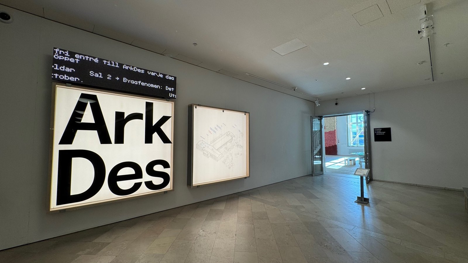 Gallery room with a large lit wall panel showing the words Ark and Des, a second white panel and an open door at the back.