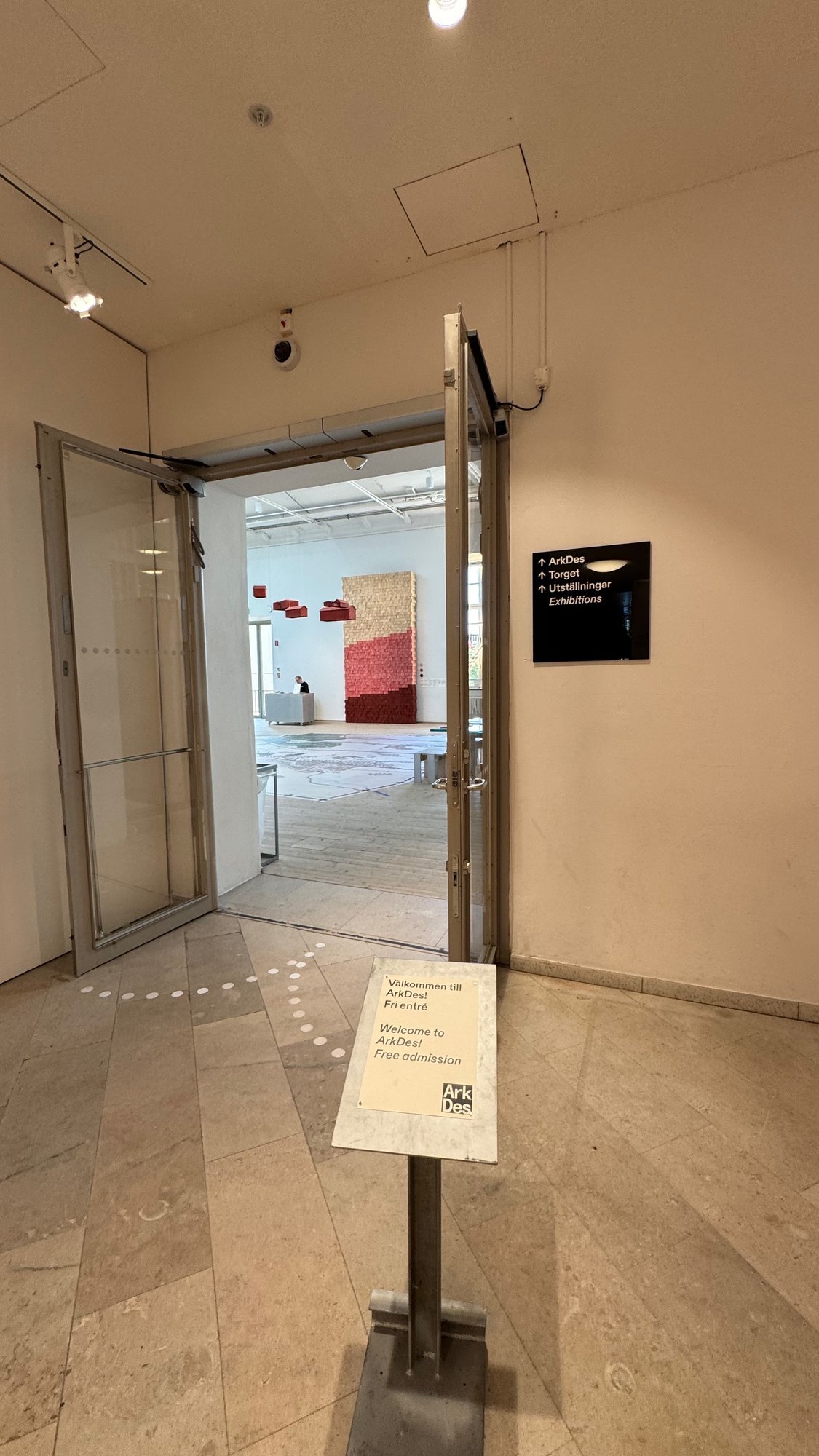 Sign on a stand beside an open glass door that says Välkommen till ArkDes! Fri entré and Welcome to ArkDes! Free admission, with a black wall sign that points to ArkDes, Torget and Utställningar, and a red and tan wall hanging in the hall behind.