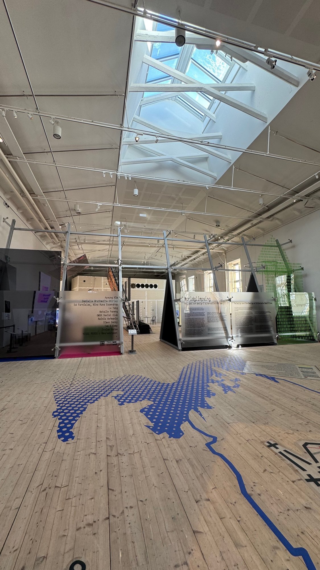 High exhibition hall with a skylight, a wooden floor with a blue map shape, and frosted glass panels with the exhibition title Worldglimpsing in Swedish and English and a list of names.