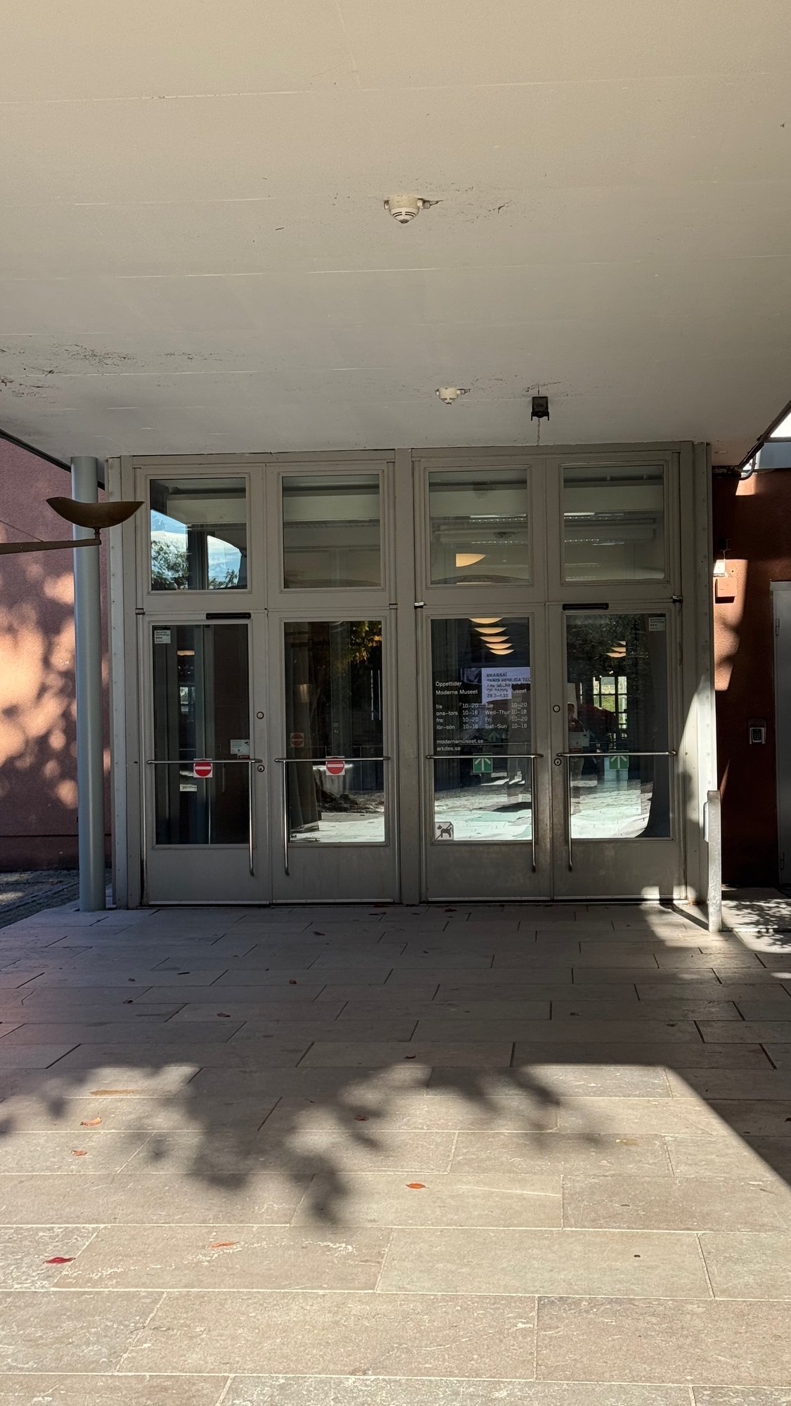Glass entrance doors under a concrete canopy, with an opening hours sign for Moderna Museet on the glass and a no dogs symbol, on a stone paved square.