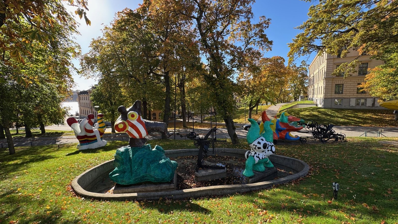 Colorful sculptures with red, white, yellow, green and blue patterns and a black metal machine in a round basin on a lawn between autumn trees, with a pale building on the right.