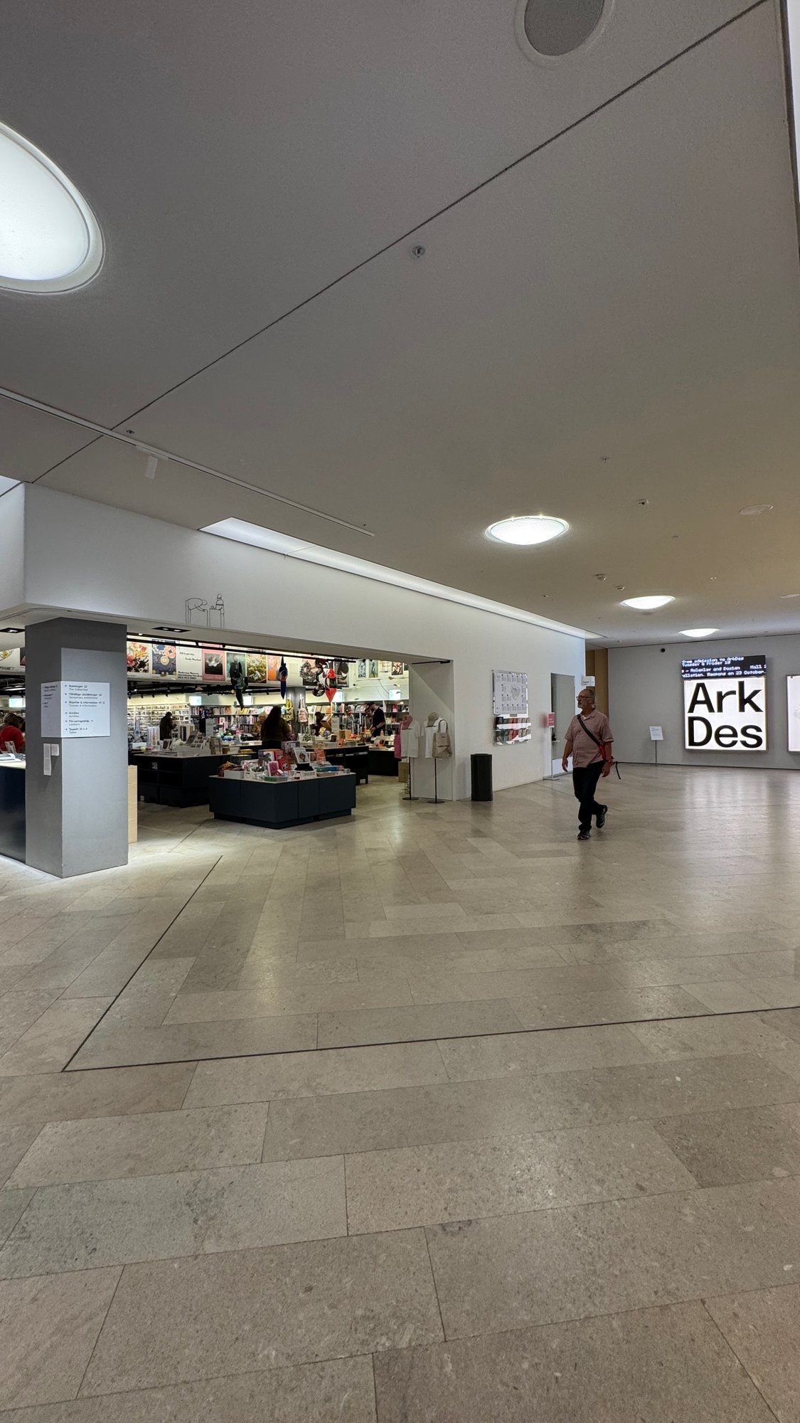 Wide stone floored hall with a museum shop with books and posters on the left and a lit wall panel with the words Ark Des at the back, with a man walking.
