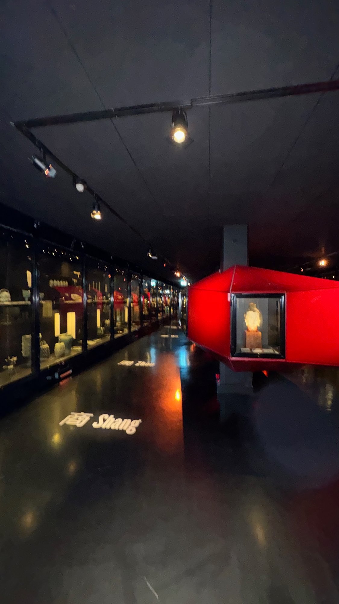 Dark gallery with lit glass cases of small objects along the left wall, a red angular display case with a sculpted head on the right, and the words Shang and Zhou in Chinese and English projected on the black floor.