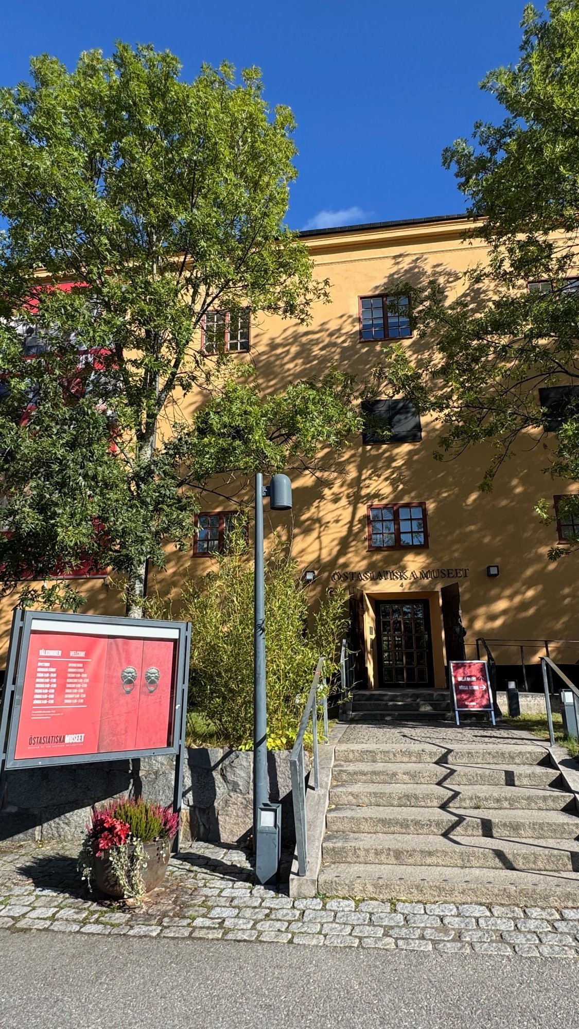 Yellow-orange museum building with the name Östasiatiska museet above a dark door, stone steps in front and a red notice board with opening hours on the left, under green trees and a blue sky.