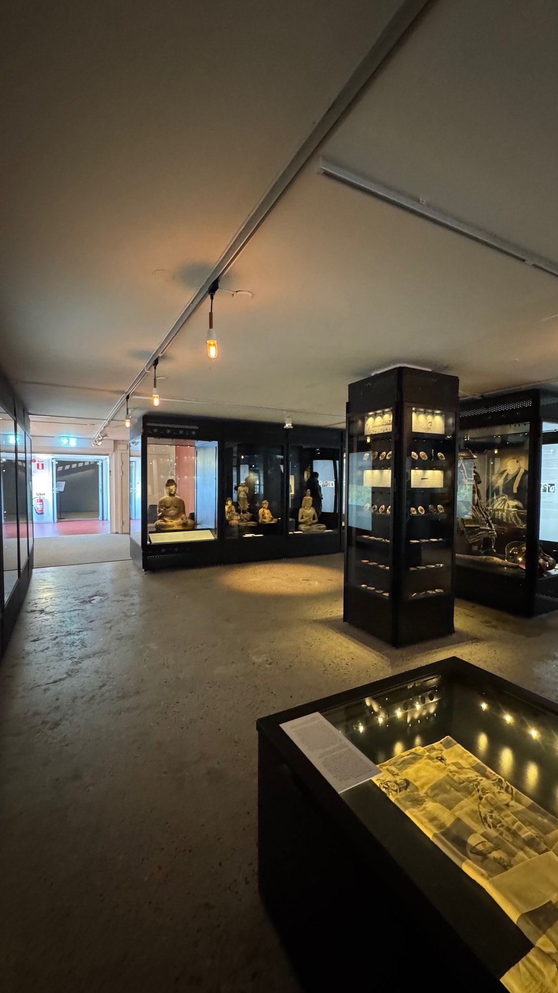 Gallery with a concrete floor and black glass cases: seated gilded Buddha figures at the back, a tall case of small round objects in the middle, a case with an armor and a portrait on the right, and a low case with a printed sheet in front.