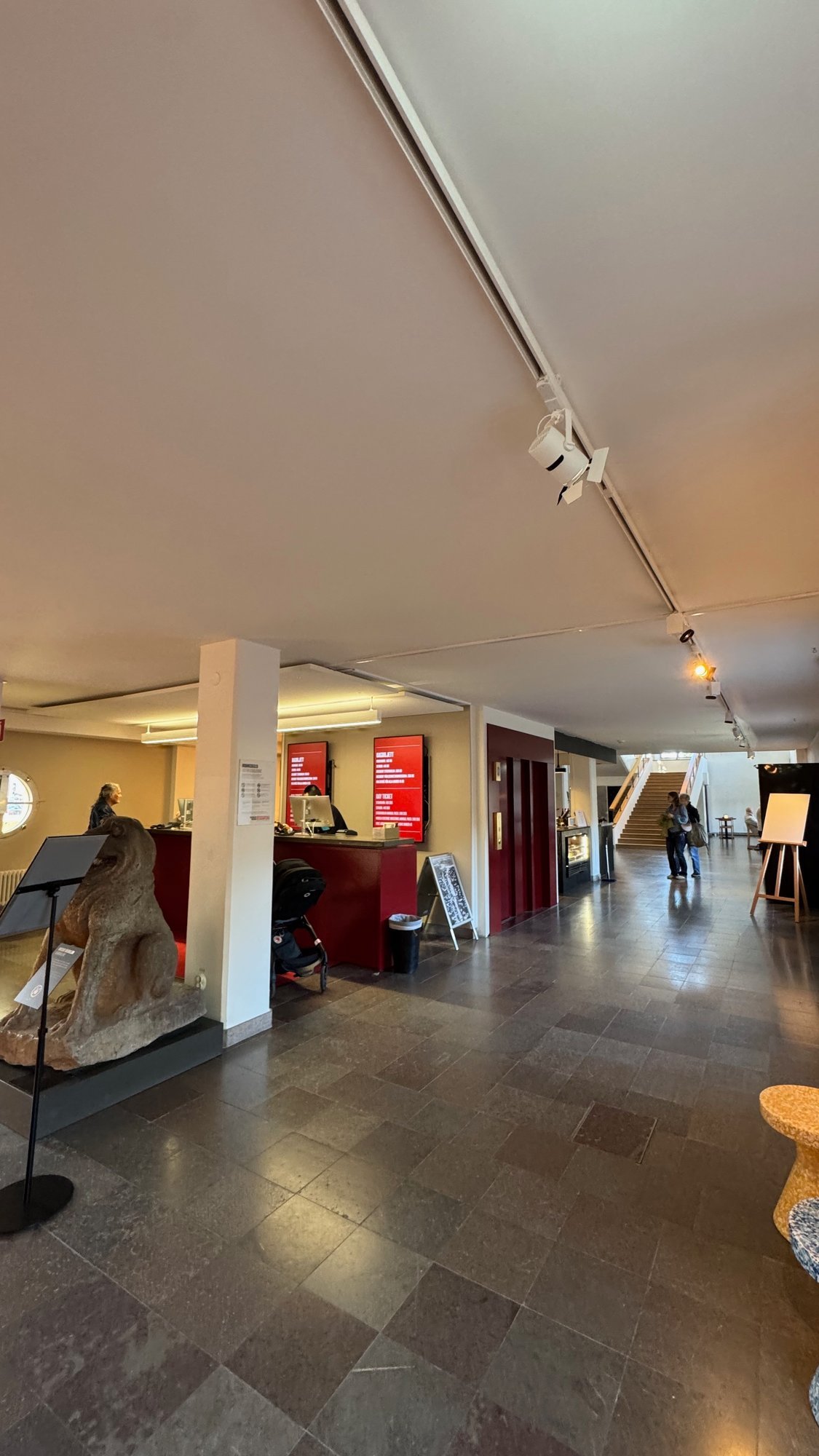 Wide entrance hall with a gray stone floor, white columns, a red reception desk with red screens, a carved stone animal on a low stand on the left and a wooden stair in the distance.