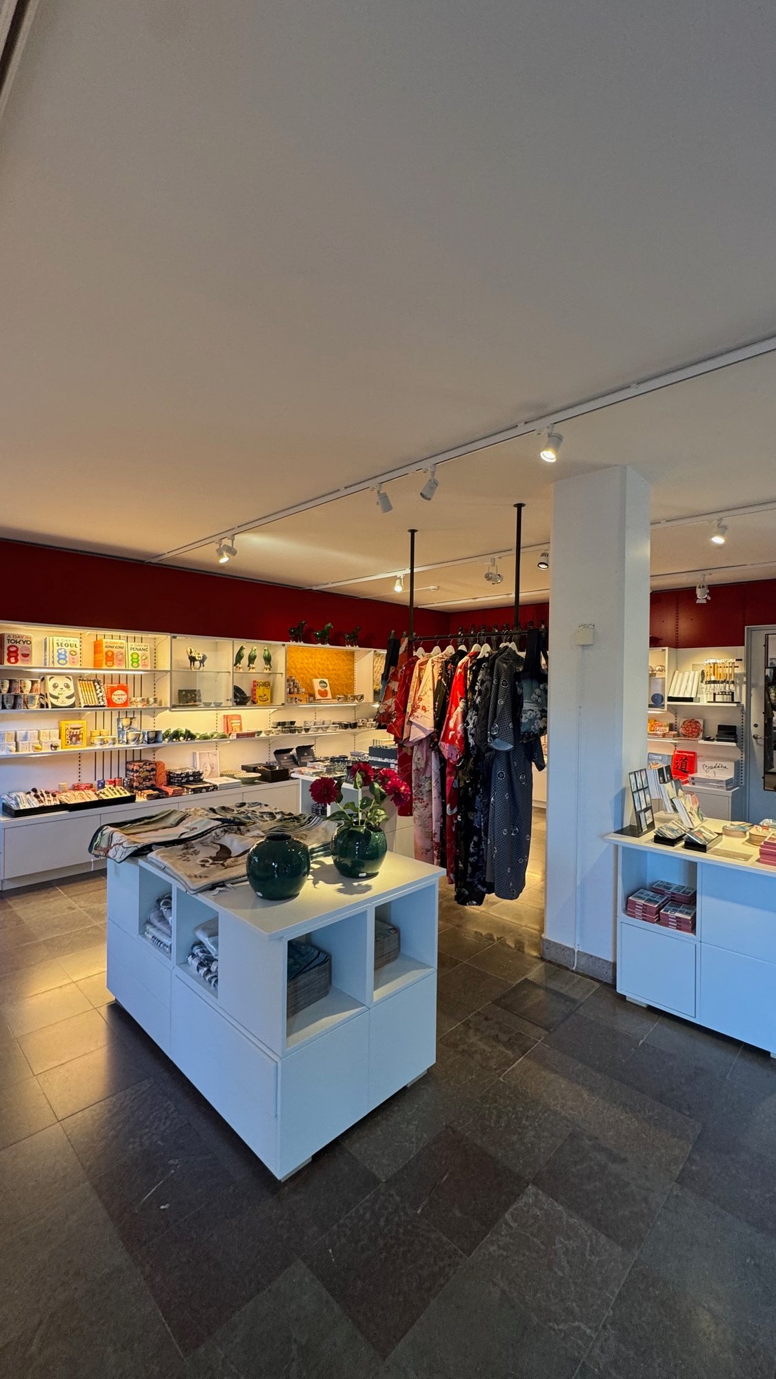 Museum shop with a red wall, white display tables and shelves with cups, books and small figures, and a rail of colorful robes.