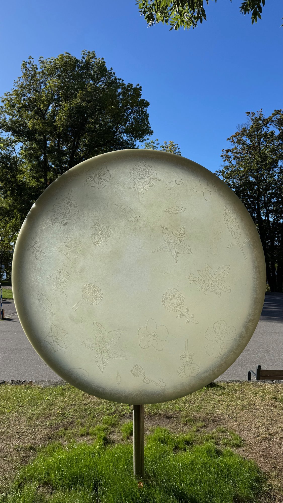 Large pale round disc on a thin pole on a lawn, with engraved drawings of flowers and leaves, in front of green trees and a blue sky.