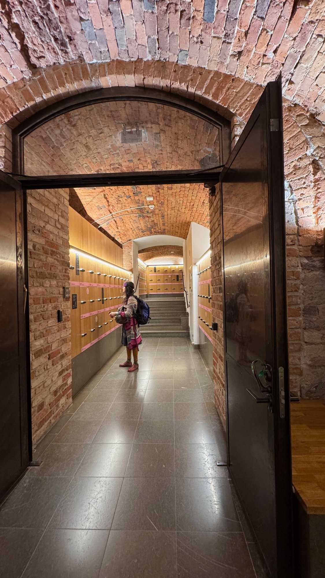 Open dark metal door in a brick arch leading to a brick-vaulted passage with wooden lockers with pink labels and a visitor standing at the lockers.