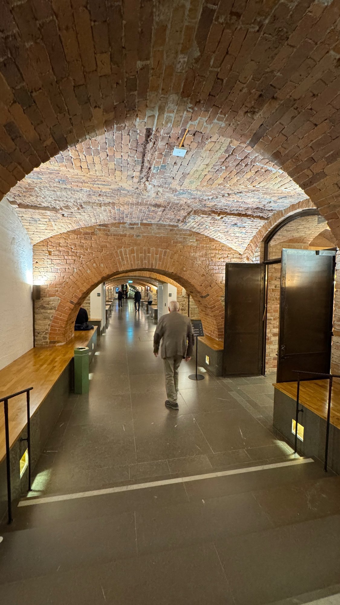 Brick-vaulted passage with several arches, wooden benches along the walls, a gray stone floor and a visitor walking away.