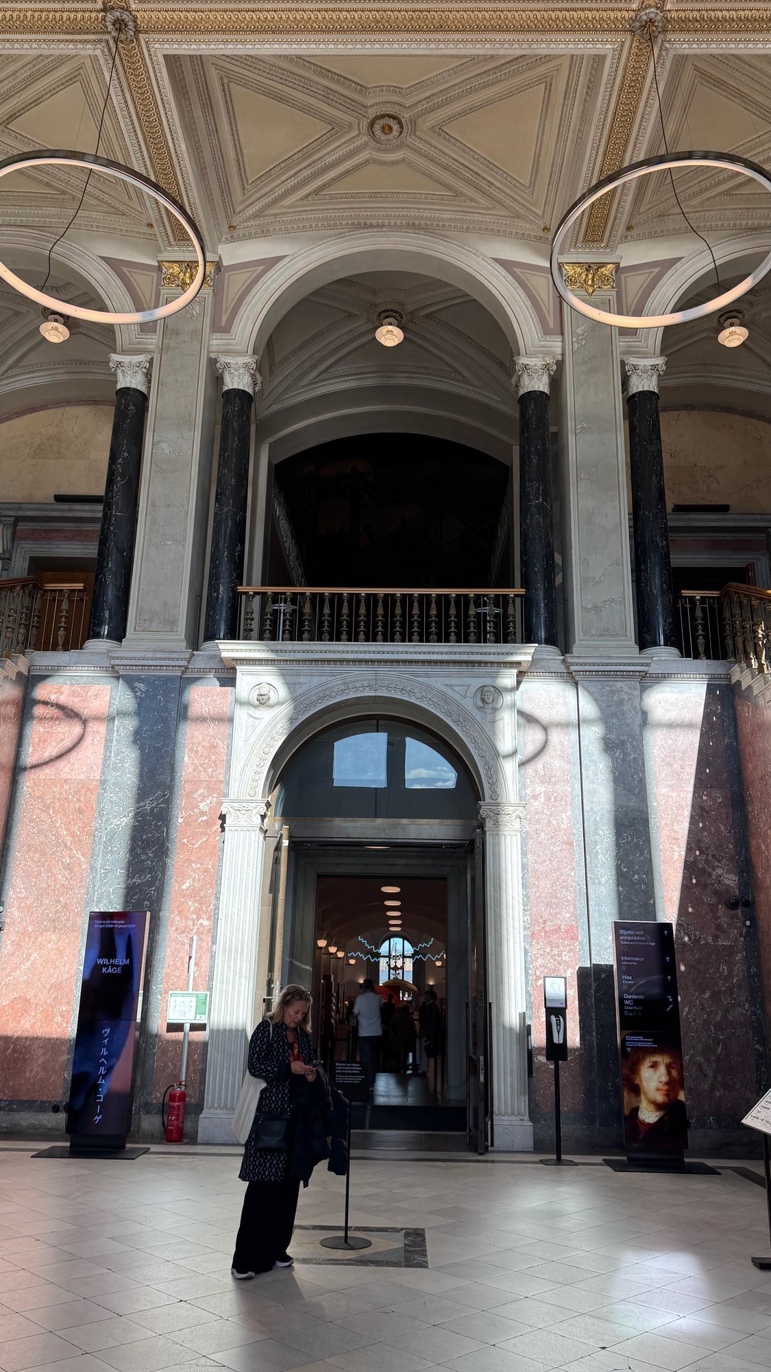 High marble hall with black columns, pink and gray marble walls, an ornate ceiling, two ring lights, a balcony above an arched doorway and signs for tickets, information, lift and cloakroom.