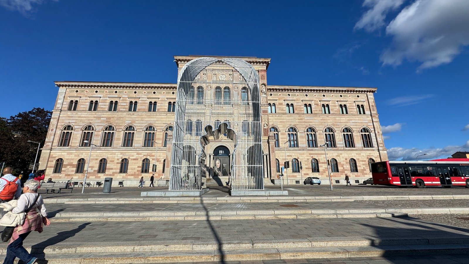 Wide view of the pale sandstone museum building with arched windows, a tall steel arch of vertical bars in front of the entrance, a red city bus on the right and a deep blue sky.