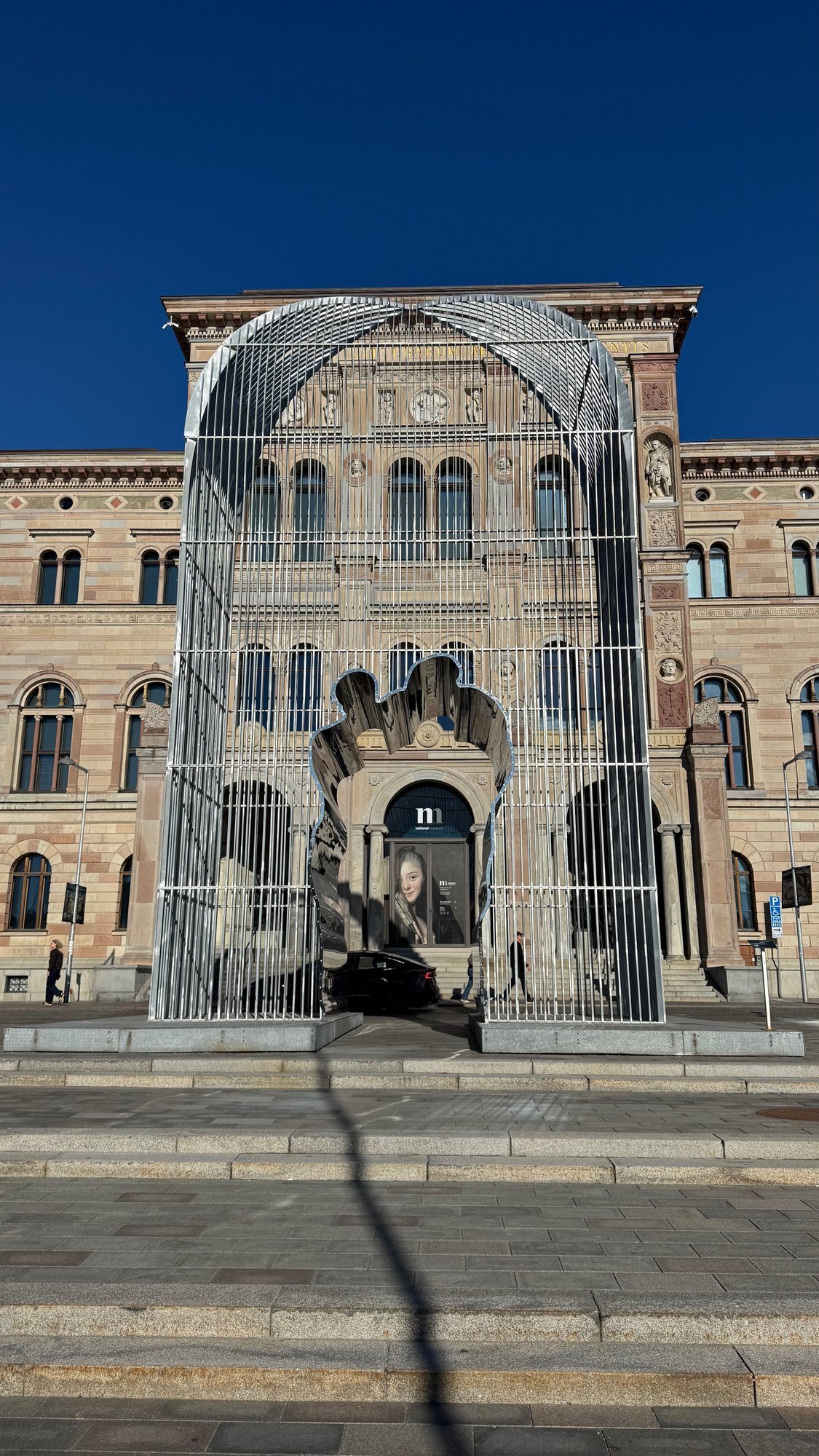 Tall steel arch of vertical bars in front of the museum entrance, with a mirror-polished silhouette of two figures inside it and the arched museum door behind.